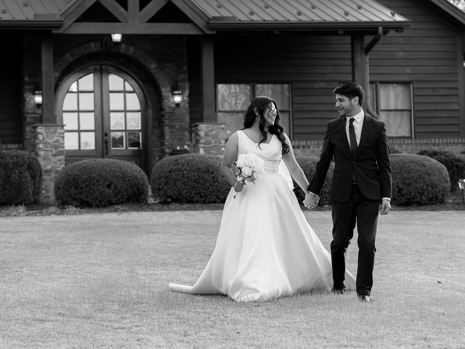 Black and white photo of a bride and groom holding hands walking outside a rustic venue on their wedding day.