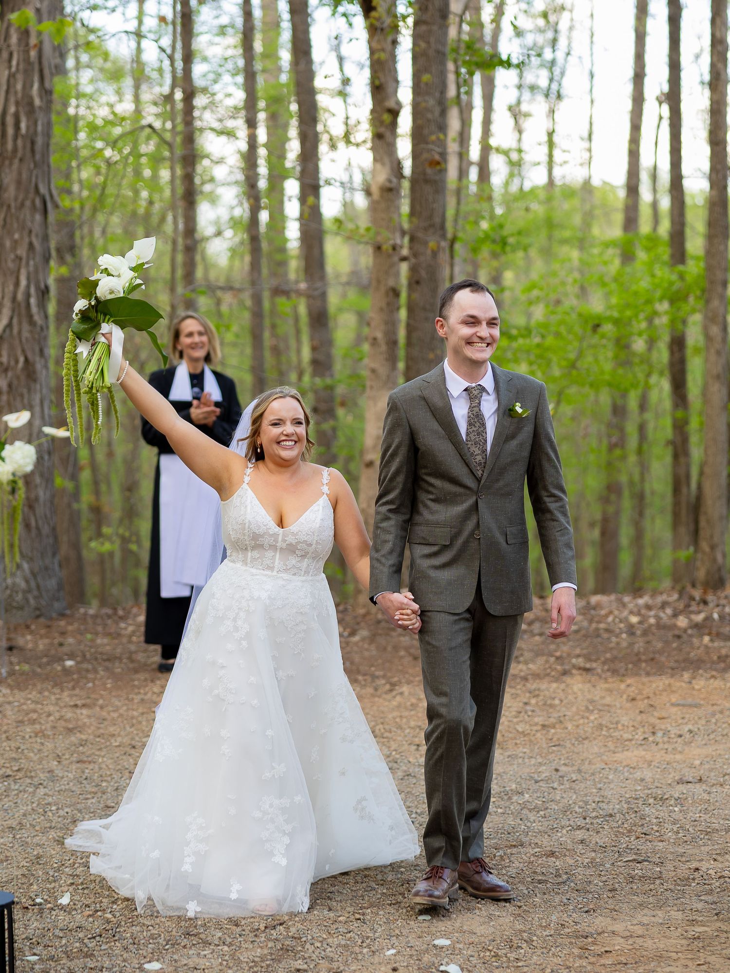 Bride raises bouquet of white flowers while holding hands with groom in gray suit, officiant behind them in wooded ceremony.