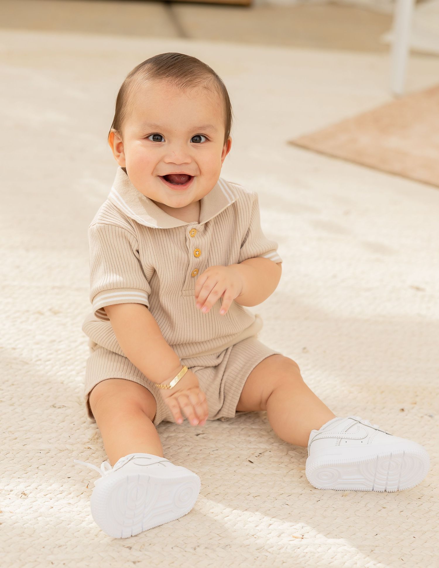 A baby in a beige ribbed romper and white shoes sits on a light carpet while playing and smiling.