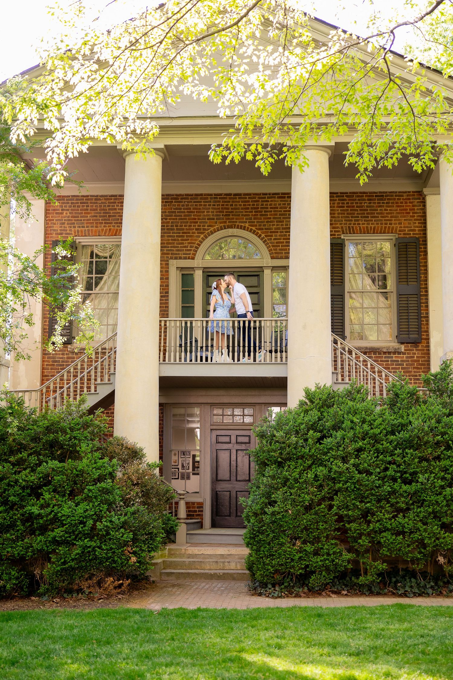 A brick house with a covered porch, surrounded by lush greenery and trees in full bloom.