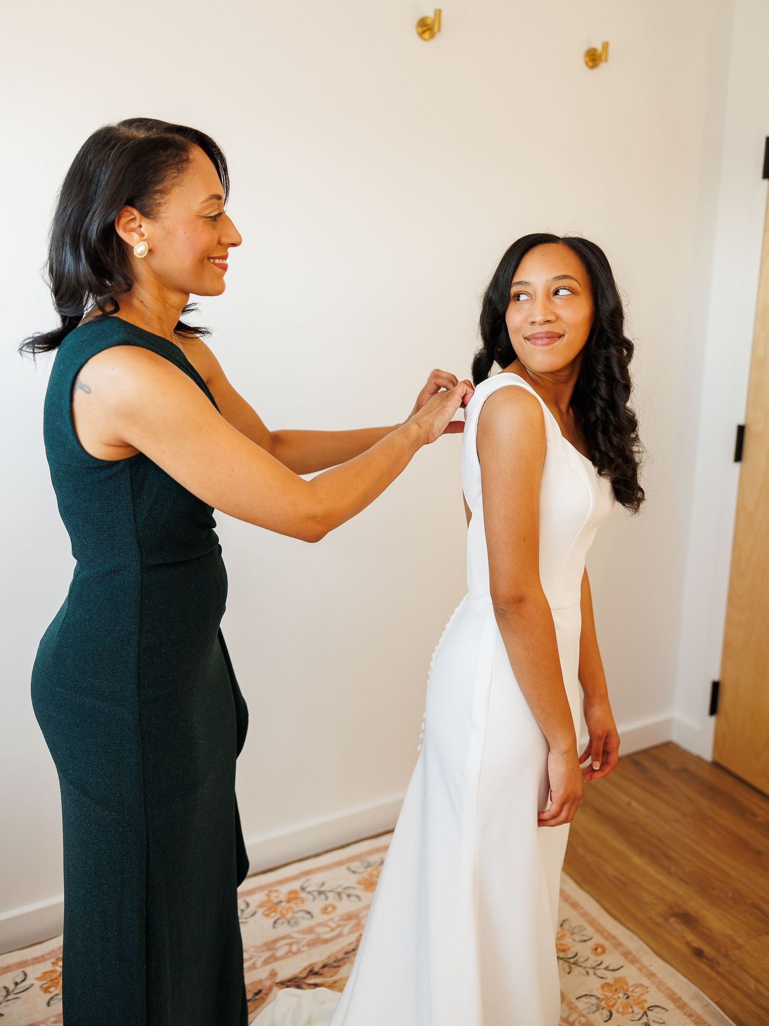 A woman in a green dress helps a bride in a white gown get dressed before her wedding ceremony.