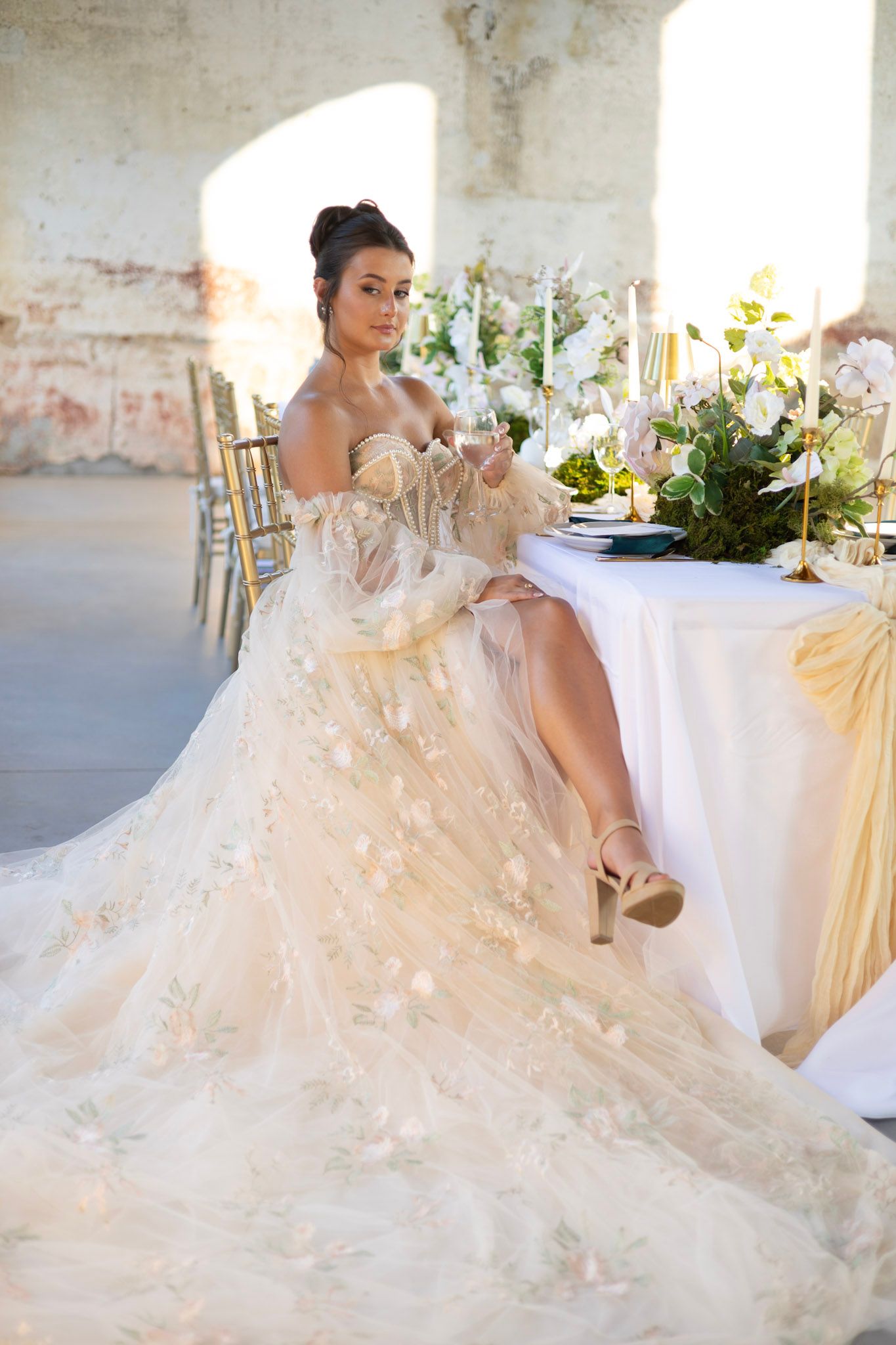 Dreamy bridal portrait in flowing white gown at elegant reception table with floral arrangements.