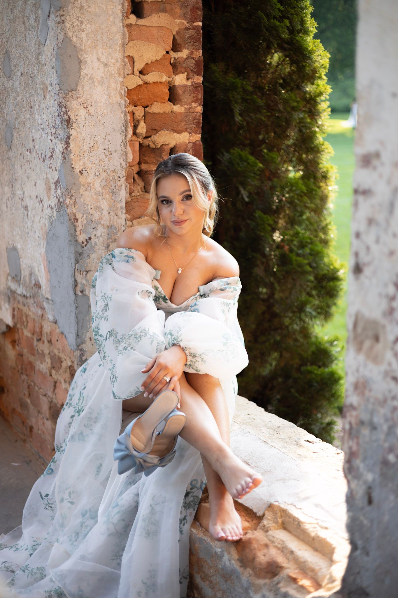 An elegant white floral wedding gown with off-shoulder design photographed against rustic stone walls and greenery.