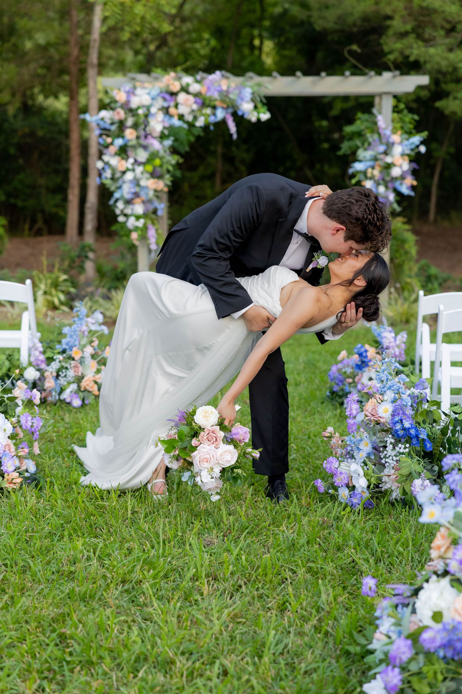 A couple in wedding attire shares a romantic dip pose among purple and blue flowers in a garden setting.