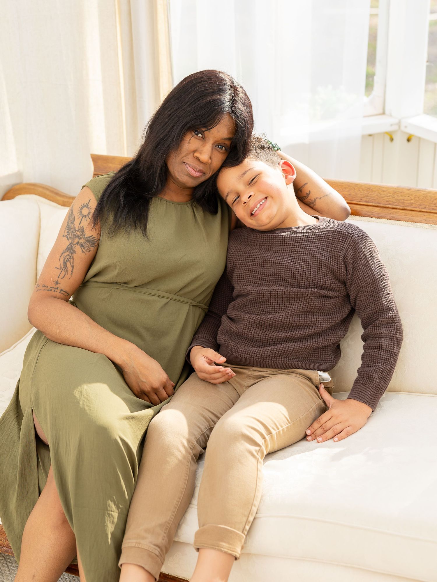 A woman in olive dress sitting on white sofa with a smiling boy in brown sweater leaning on her.