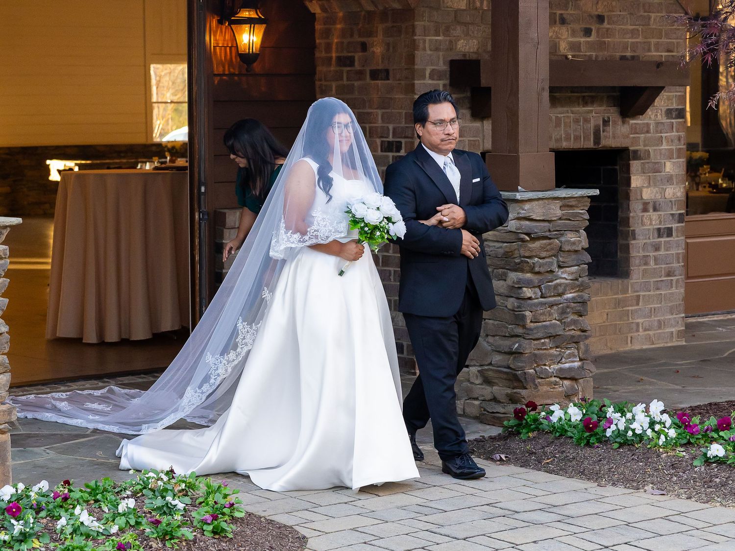 Bride in white gown with long lace veil walks with escort outside stone wedding venue.