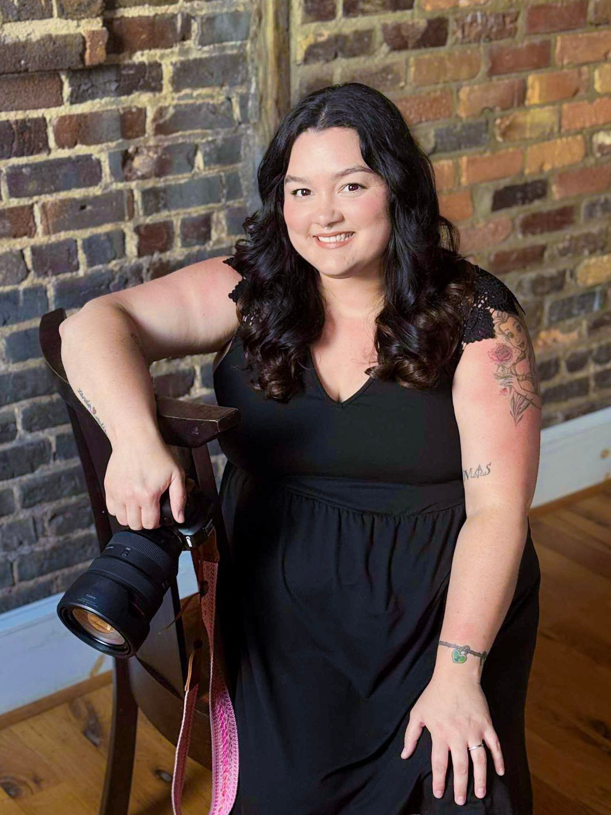 A photographer in a black dress sitting with camera equipment against a brick wall.