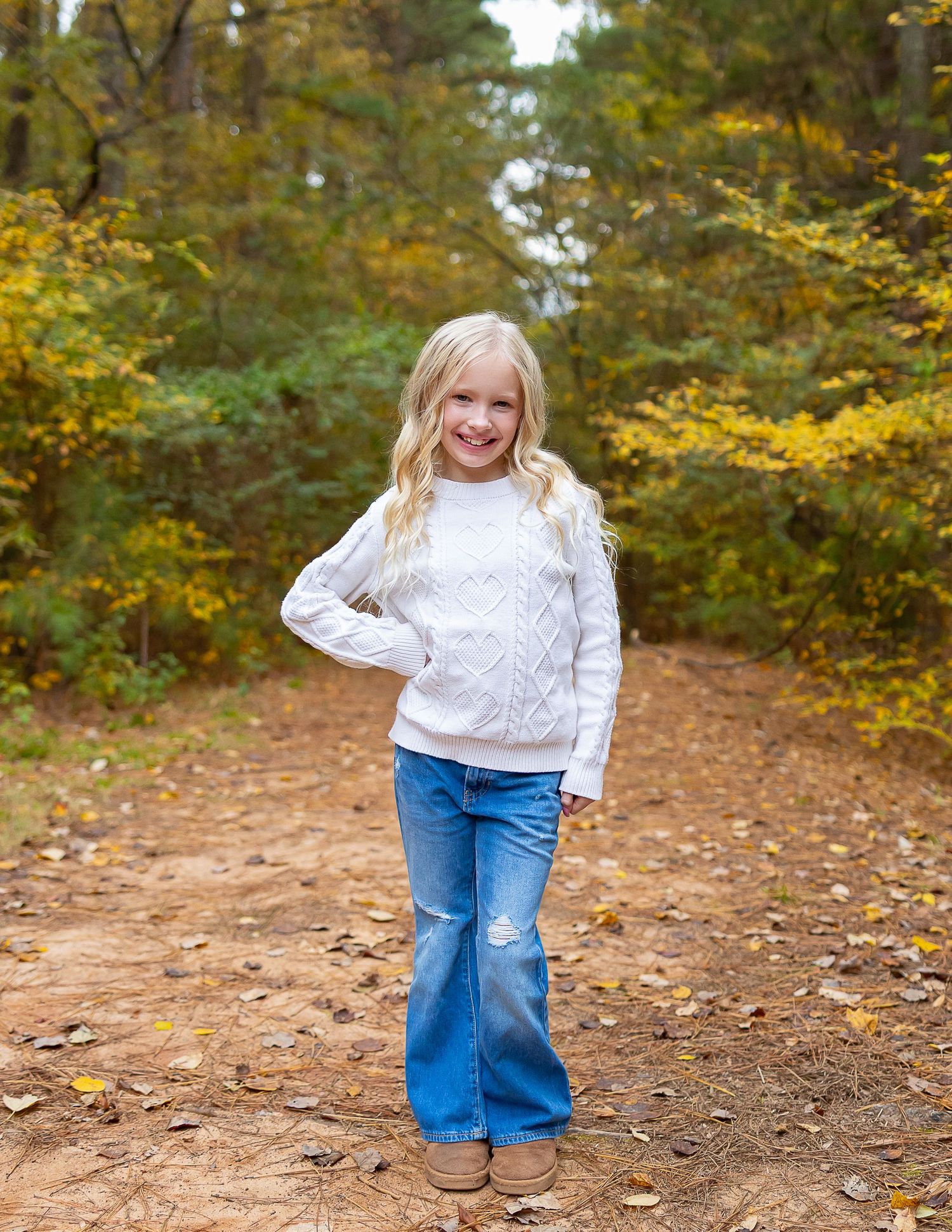 A child in a white cable knit sweater and jeans poses on a forest path surrounded by autumn foliage.