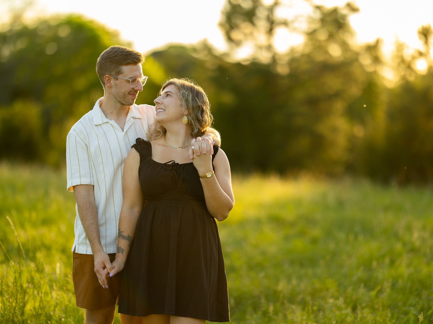 Couple holding hands smiling at each other in a sunny green field with golden hour light and trees in background.