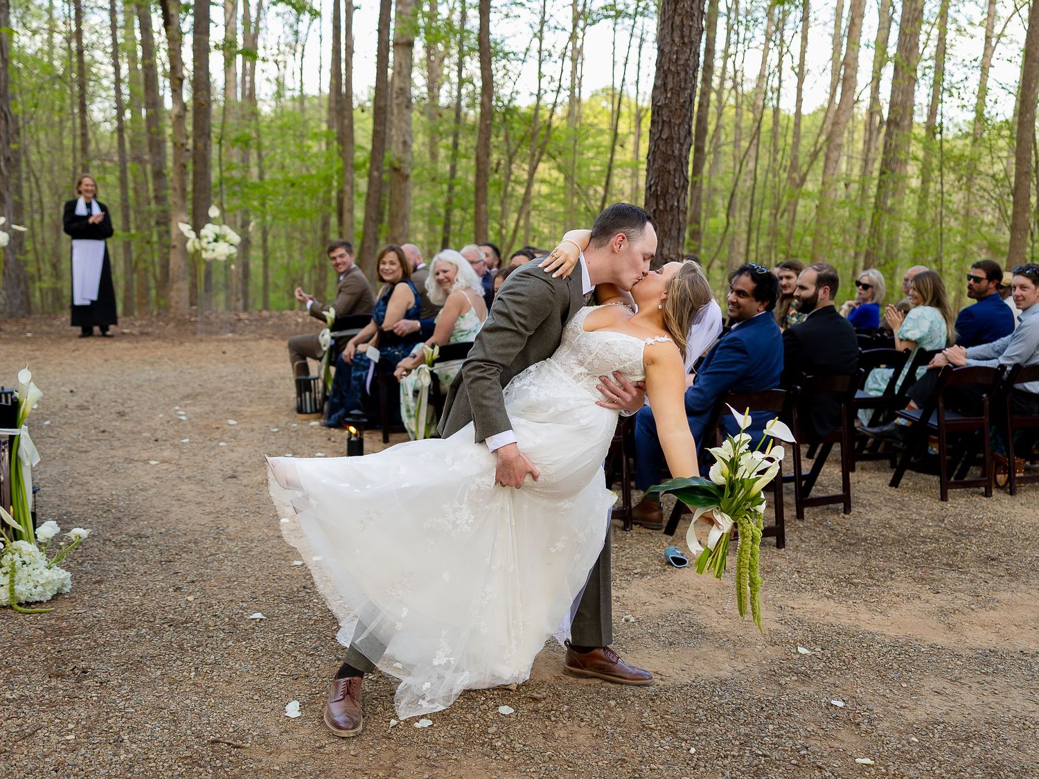 Bride and groom share a romantic dip kiss during outdoor wedding ceremony in a pine forest with guests seated behind.
