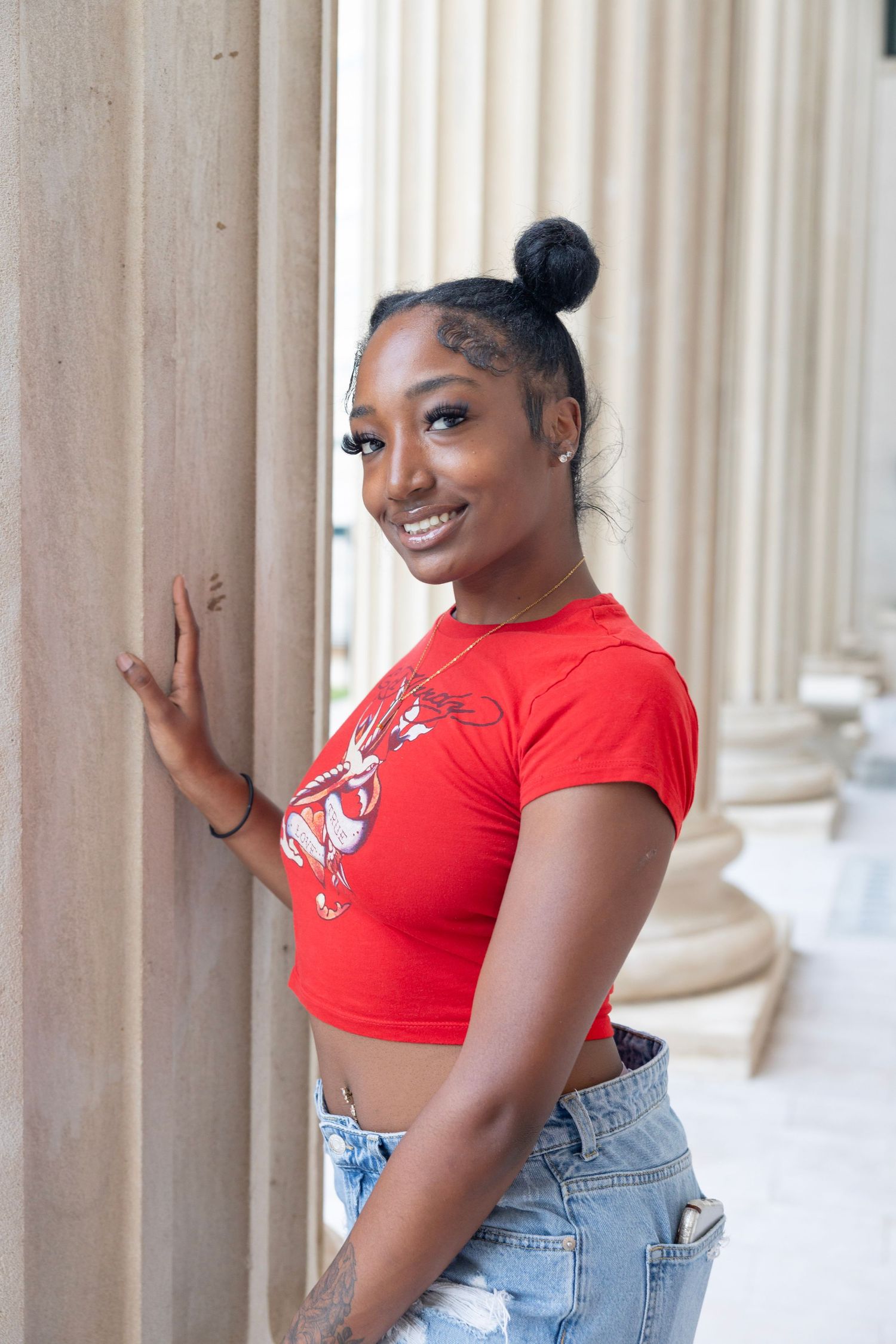 Young woman in red crop top and ripped jeans poses cheerfully against classical stone column background.