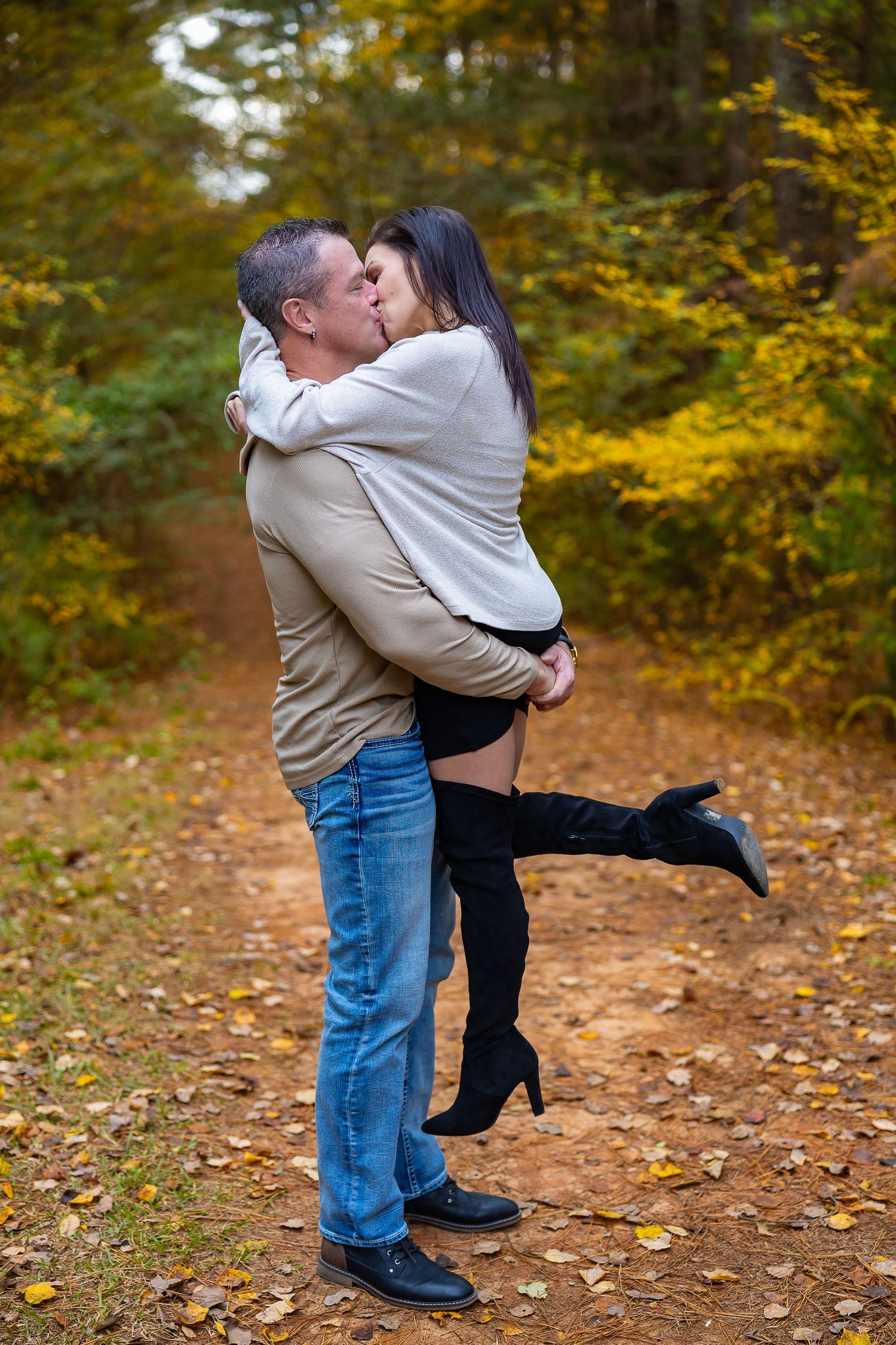 A romantic couple embraces and kisses on a path covered in autumn leaves during a fall photo session.