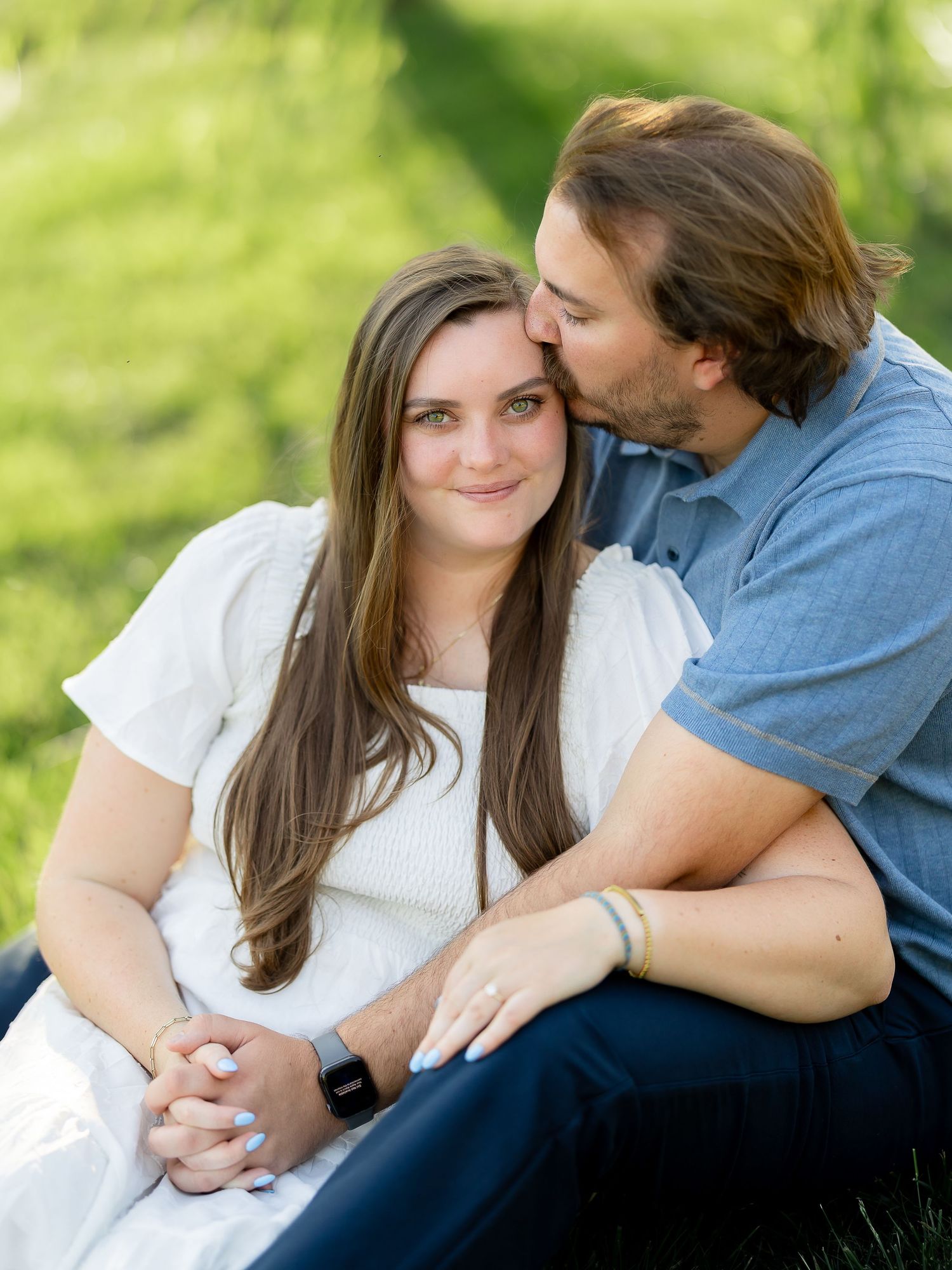 Couple sitting in green grass, man kissing woman on forehead during outdoor engagement photo session.
