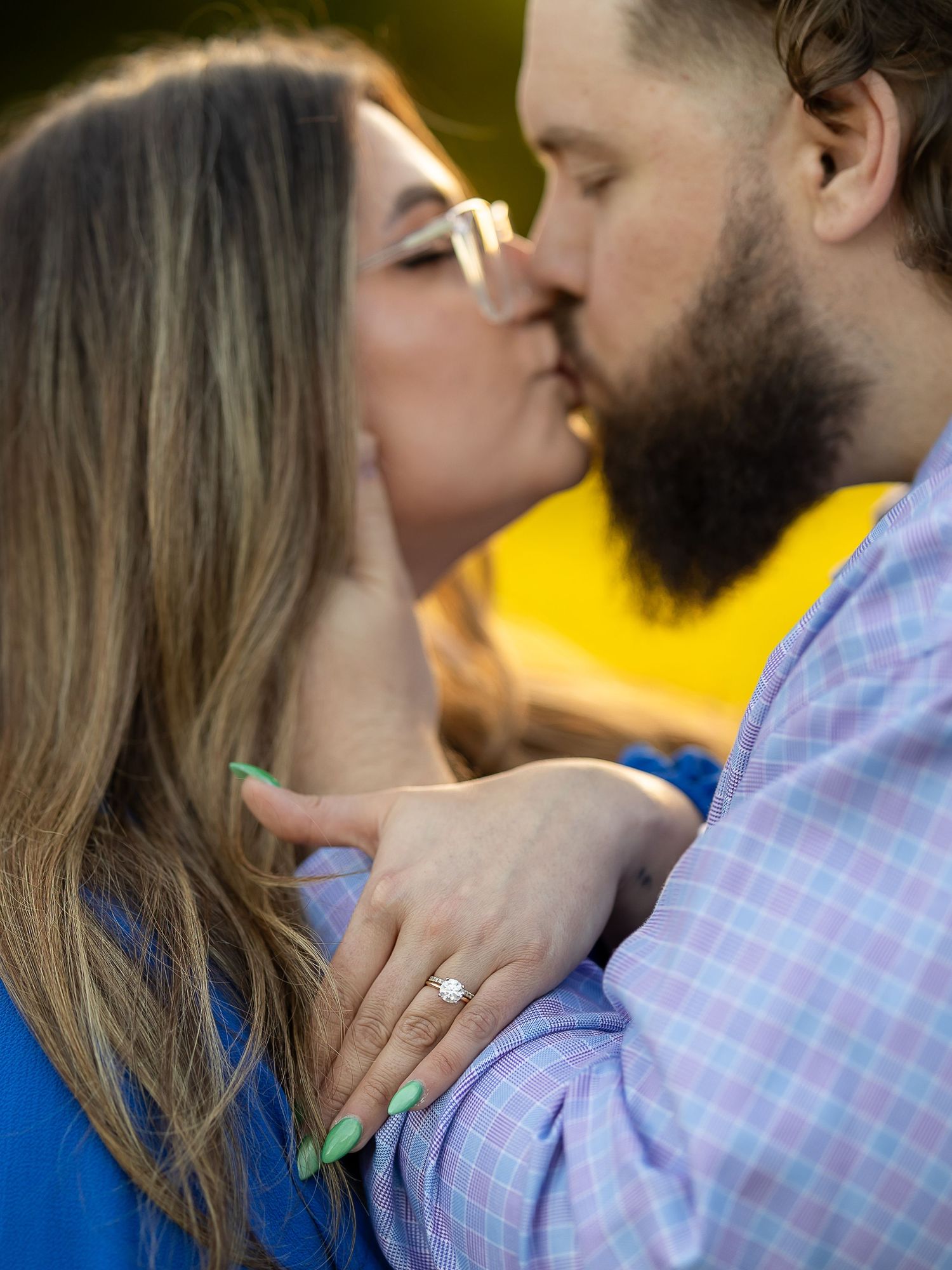 A couple shares a romantic kiss during an engagement photo session, showcasing a sparkling diamond ring with green manicured nails.
