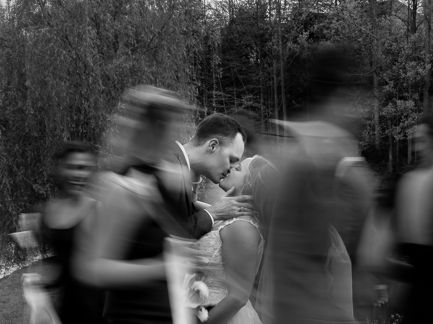 Black and white wedding photo of couple kissing while blurred guests move around them outdoors.