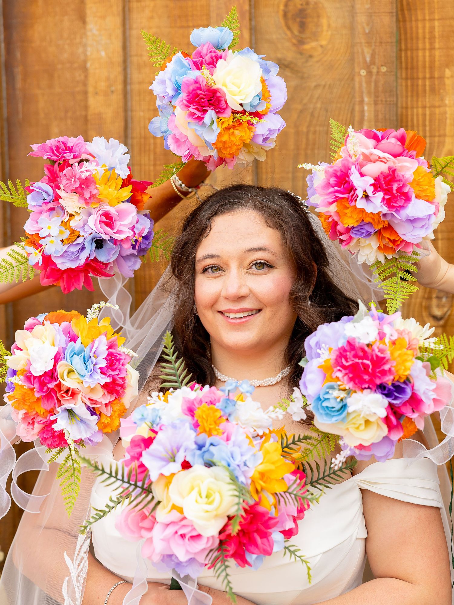 Smiling bride in white dress and veil surrounded by colorful rainbow floral bouquets on her wedding day.