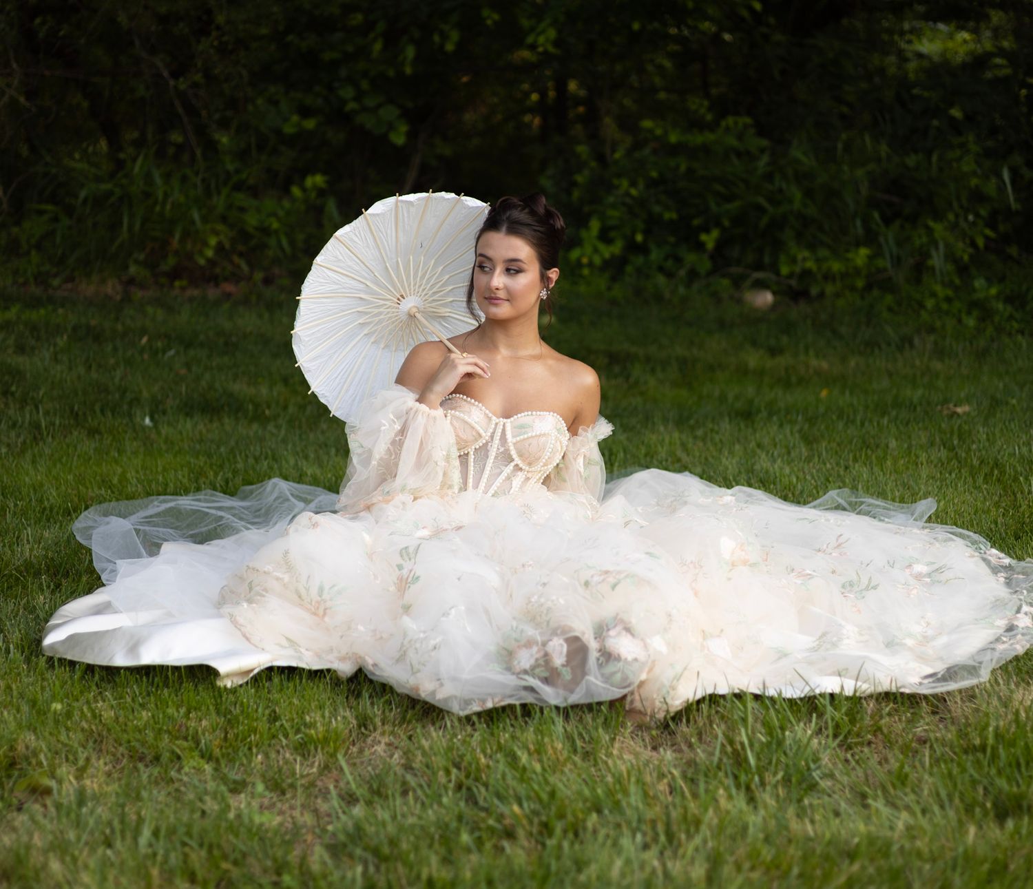 A bride in a flowing white wedding gown lies gracefully on green grass, her dress billowing around her in an artistic pose.