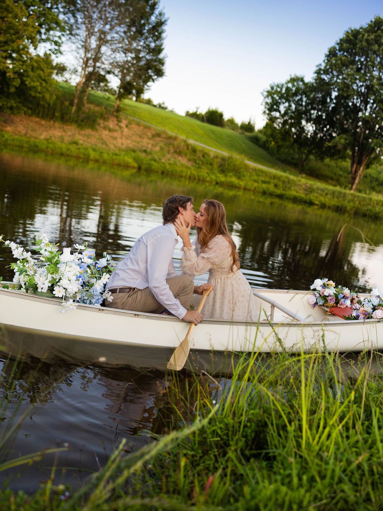 A romantic couple shares an intimate moment on a wooden dock by a serene lake surrounded by lush greenery at sunset.