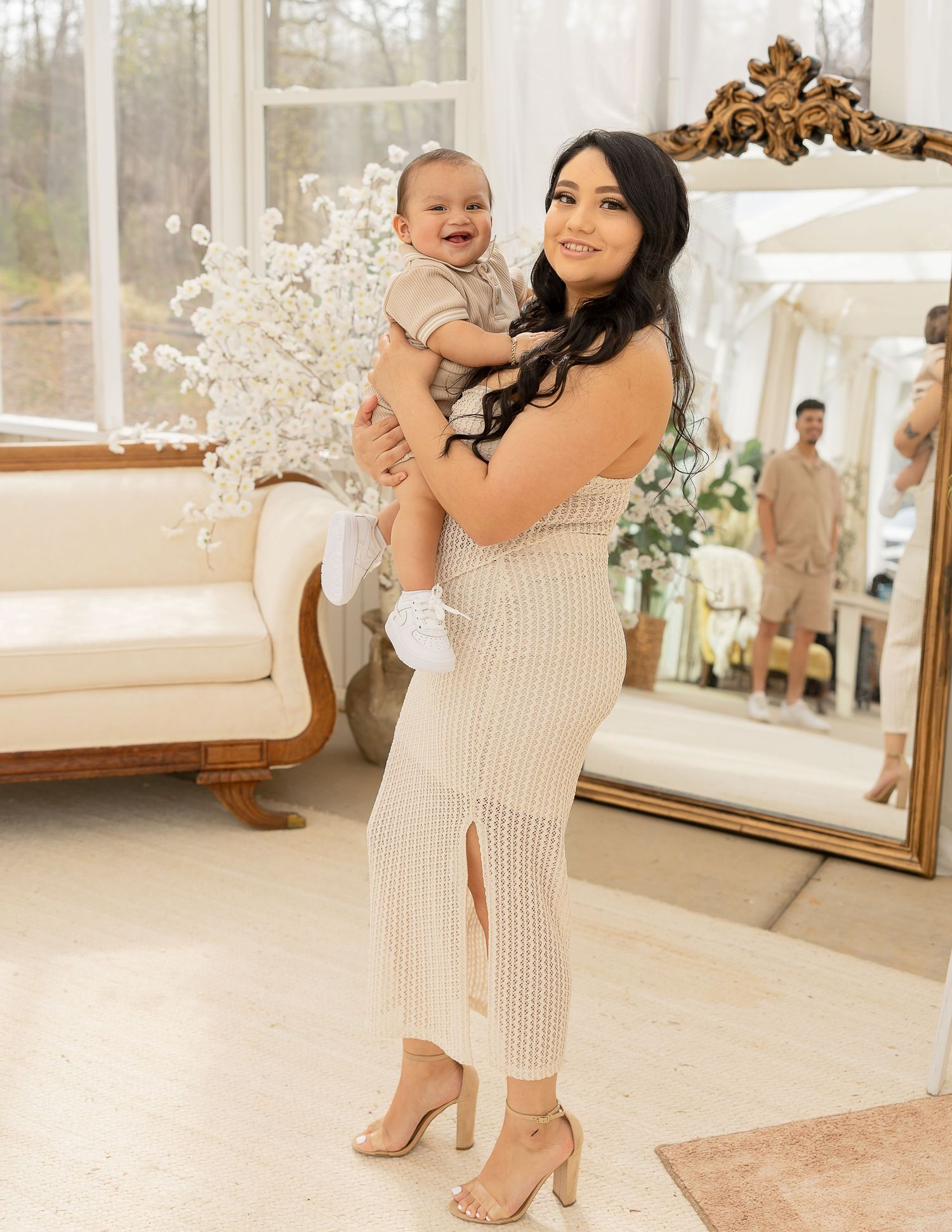 A parent and baby pose together in an elegant room with a decorative mirror and floral decor.