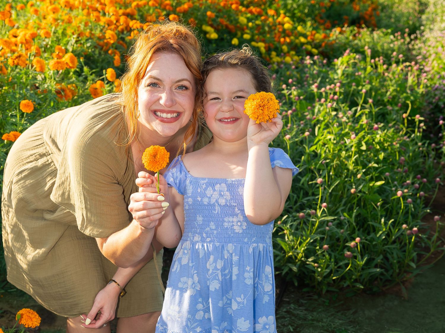 Woman and young girl smiling in a vibrant orange flower garden, each holding a marigold bloom outdoors.