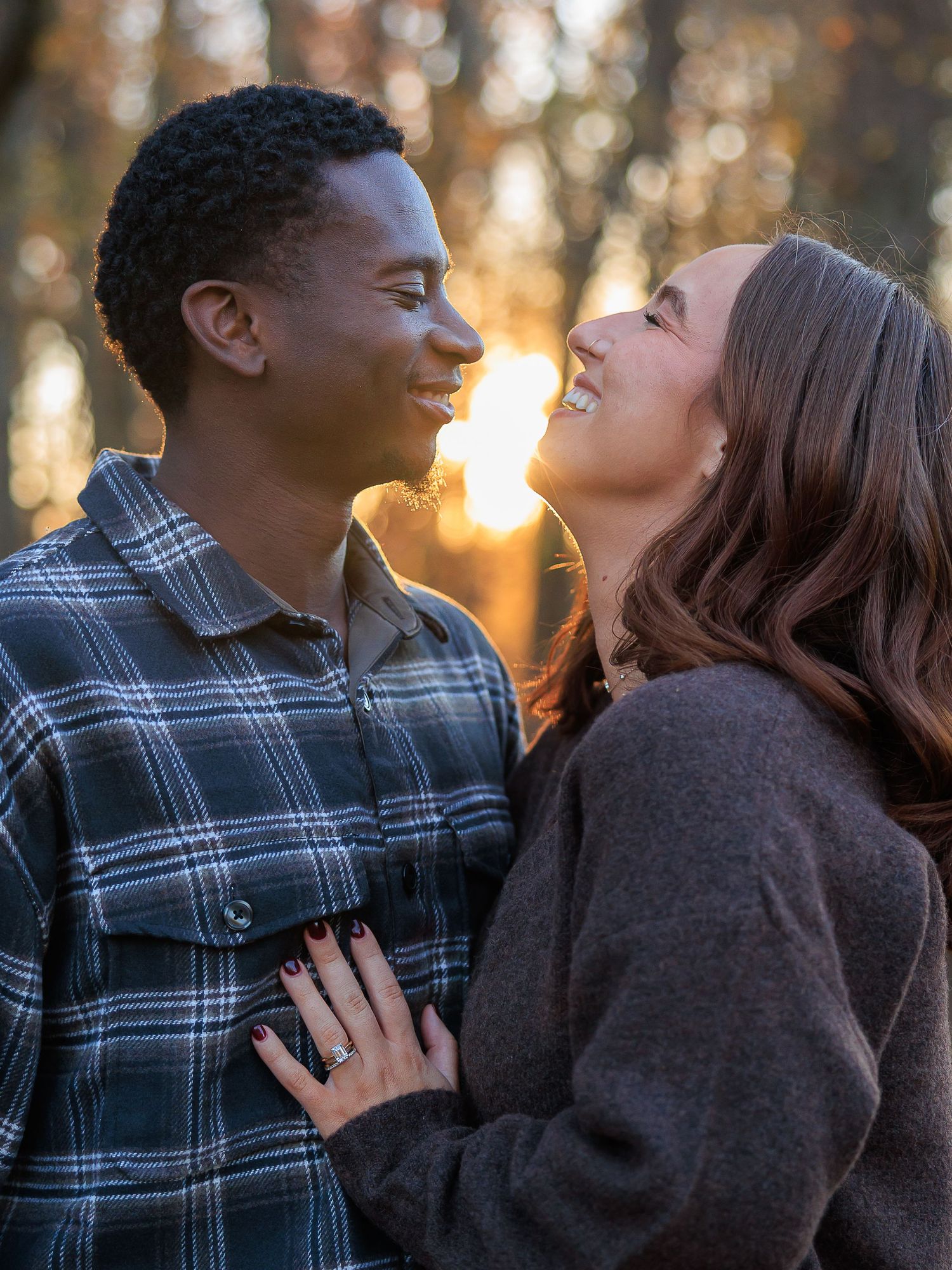 A couple shares romantic moments in a sunset-lit forest setting wearing plaid and dark sweaters.