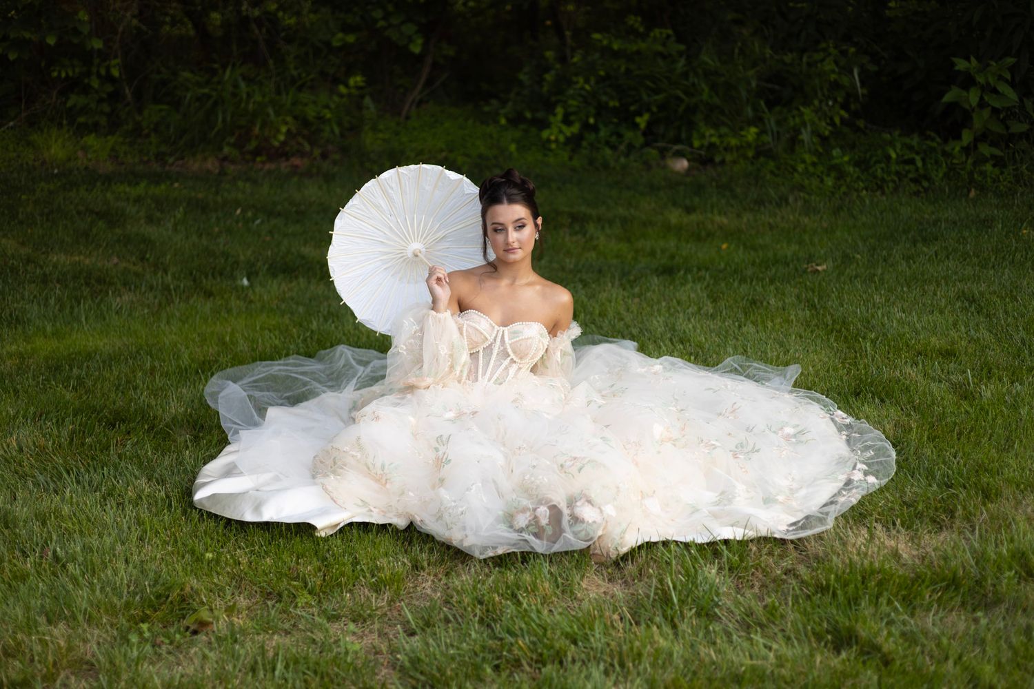A bride in a flowing white wedding dress sits on grass while holding a white parasol on a summer day.