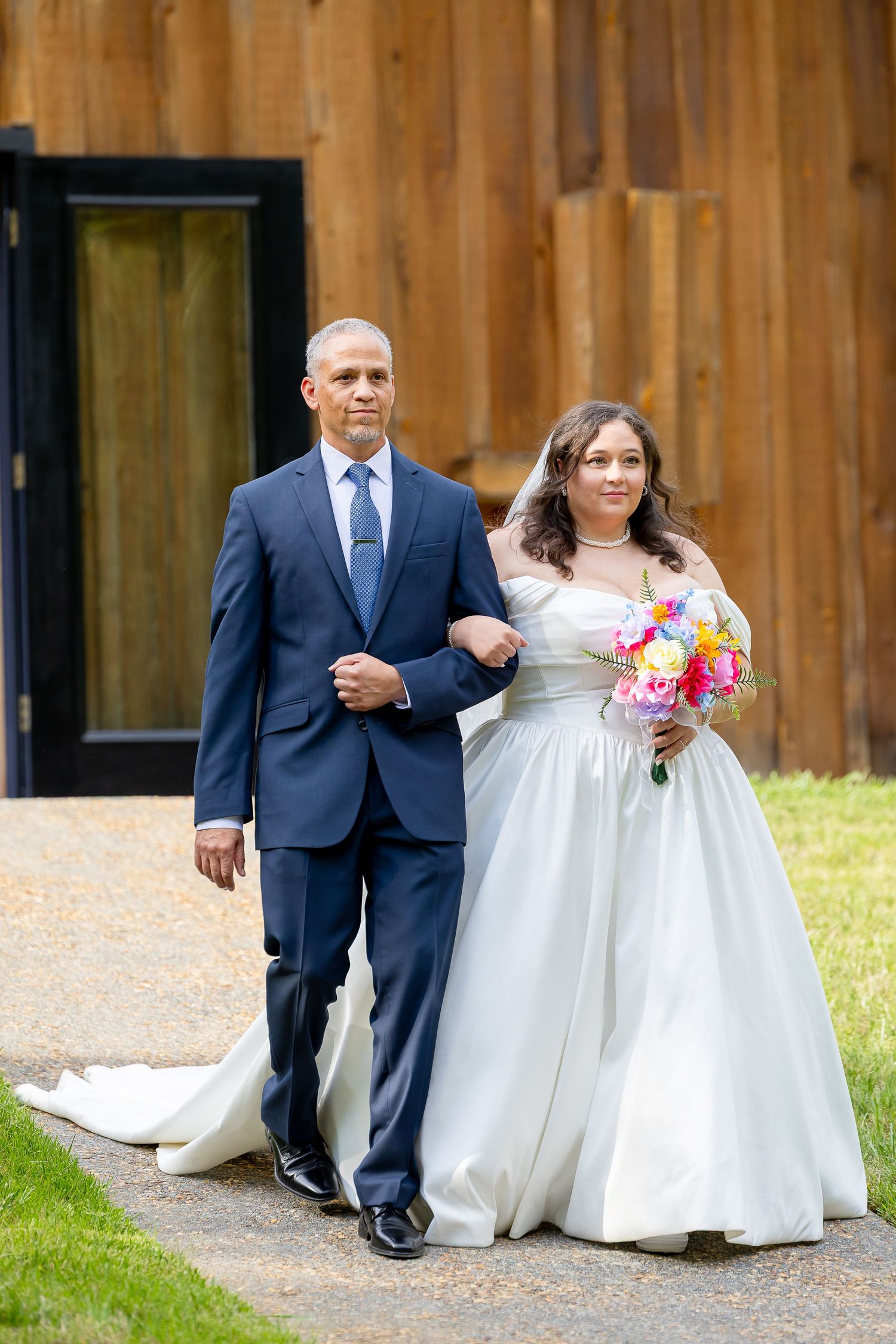 Bride in white ball gown holding colorful bouquet walks arm-in-arm with man in navy suit on garden path.