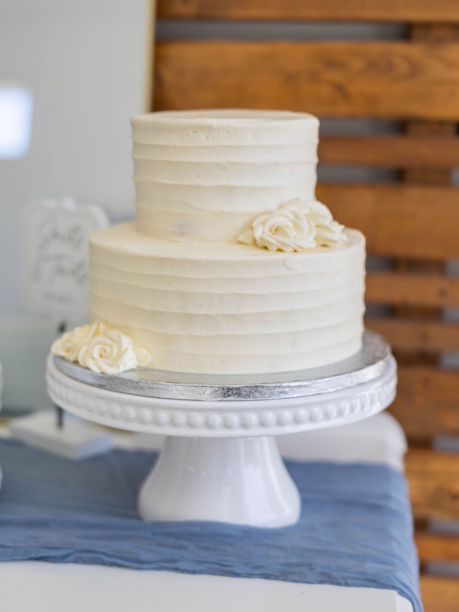 Elegant two-tier white wedding cake with textured buttercream frosting and ivory roses on a white pedestal stand.