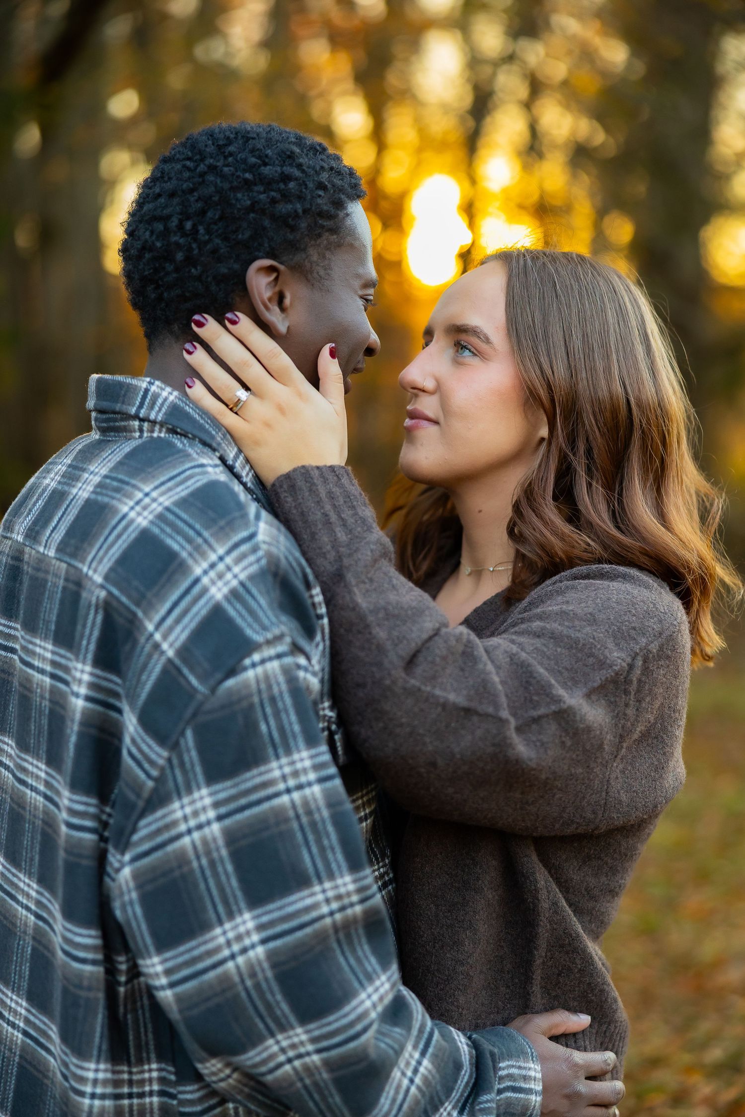 A couple embraces tenderly in an autumn forest setting with golden sunlight filtering through the trees in the background.