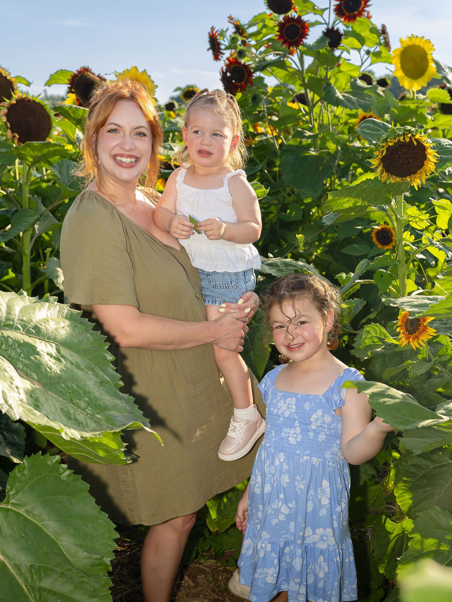 A woman in an olive dress holds a toddler while a young girl in blue stands beside them in a sunflower field.