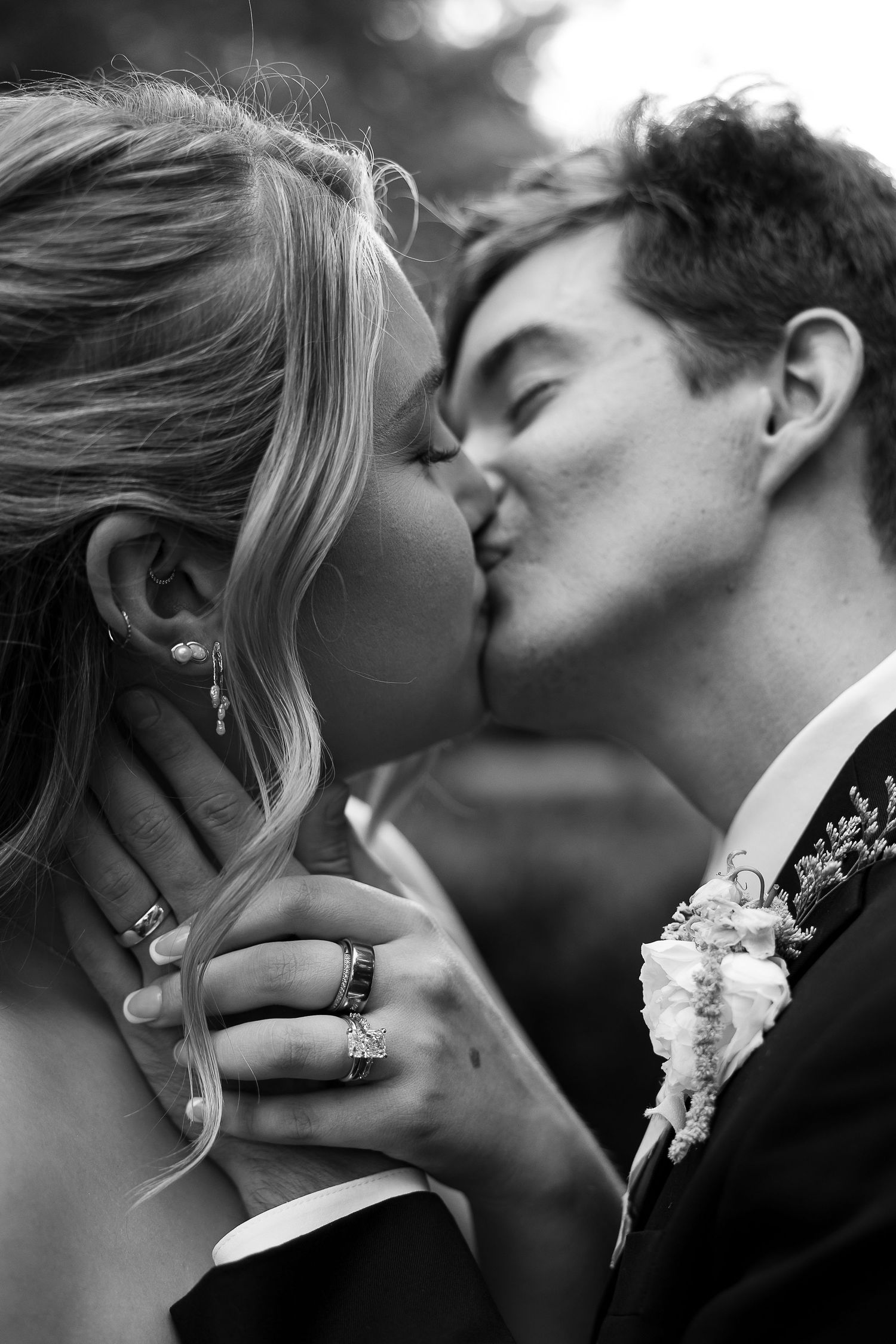 Black and white photo of a couple sharing a romantic kiss, bride wearing rings, groom in suit with boutonniere.