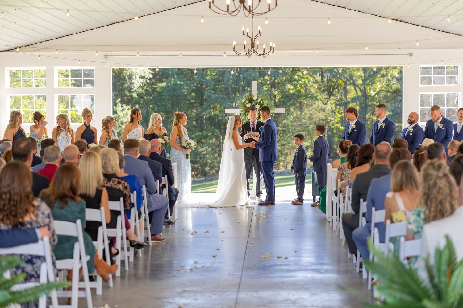 Elegant outdoor wedding ceremony under a white tent with guests seated in white chairs along a floral petal aisle.
