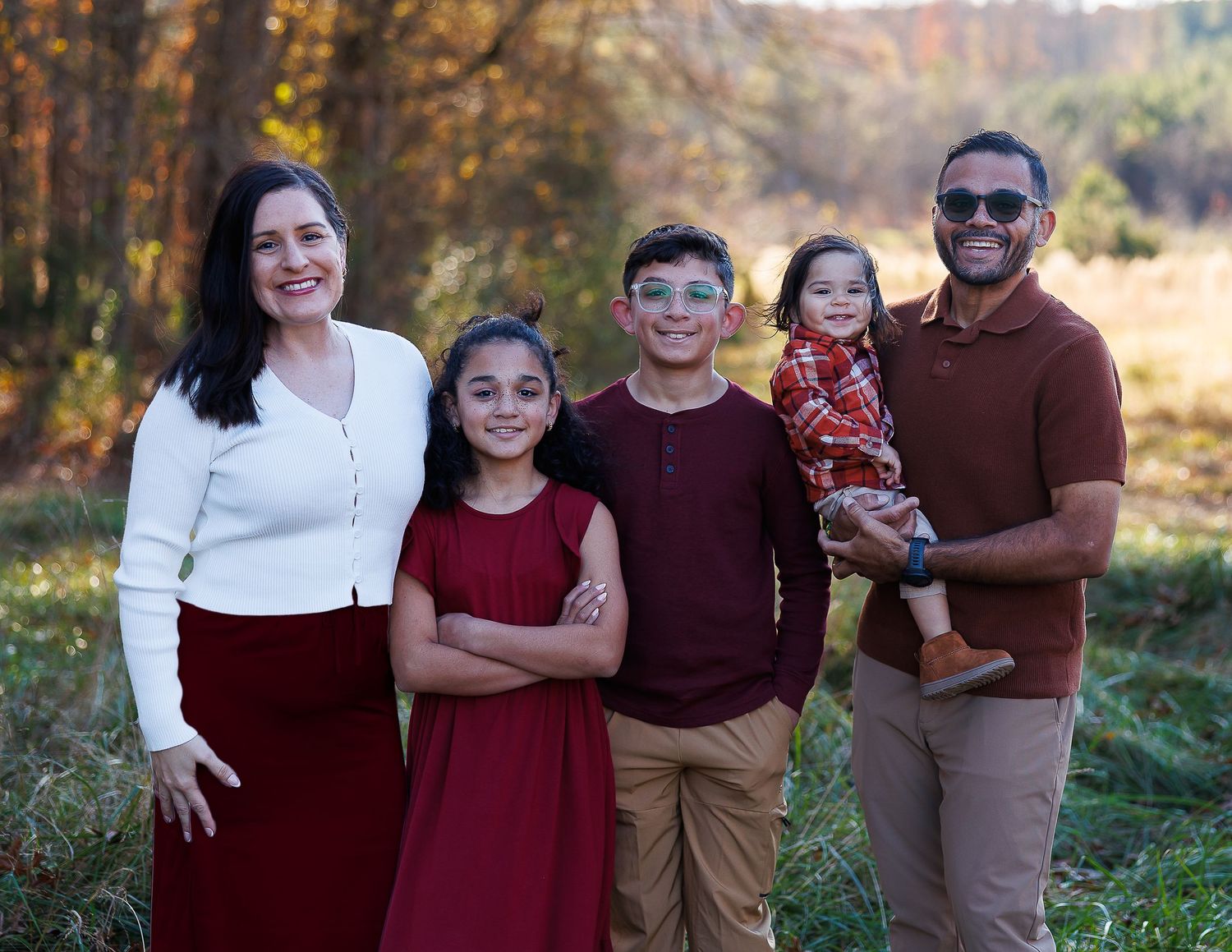 A family group wearing coordinated burgundy and white clothing stands together in an autumn forest setting.