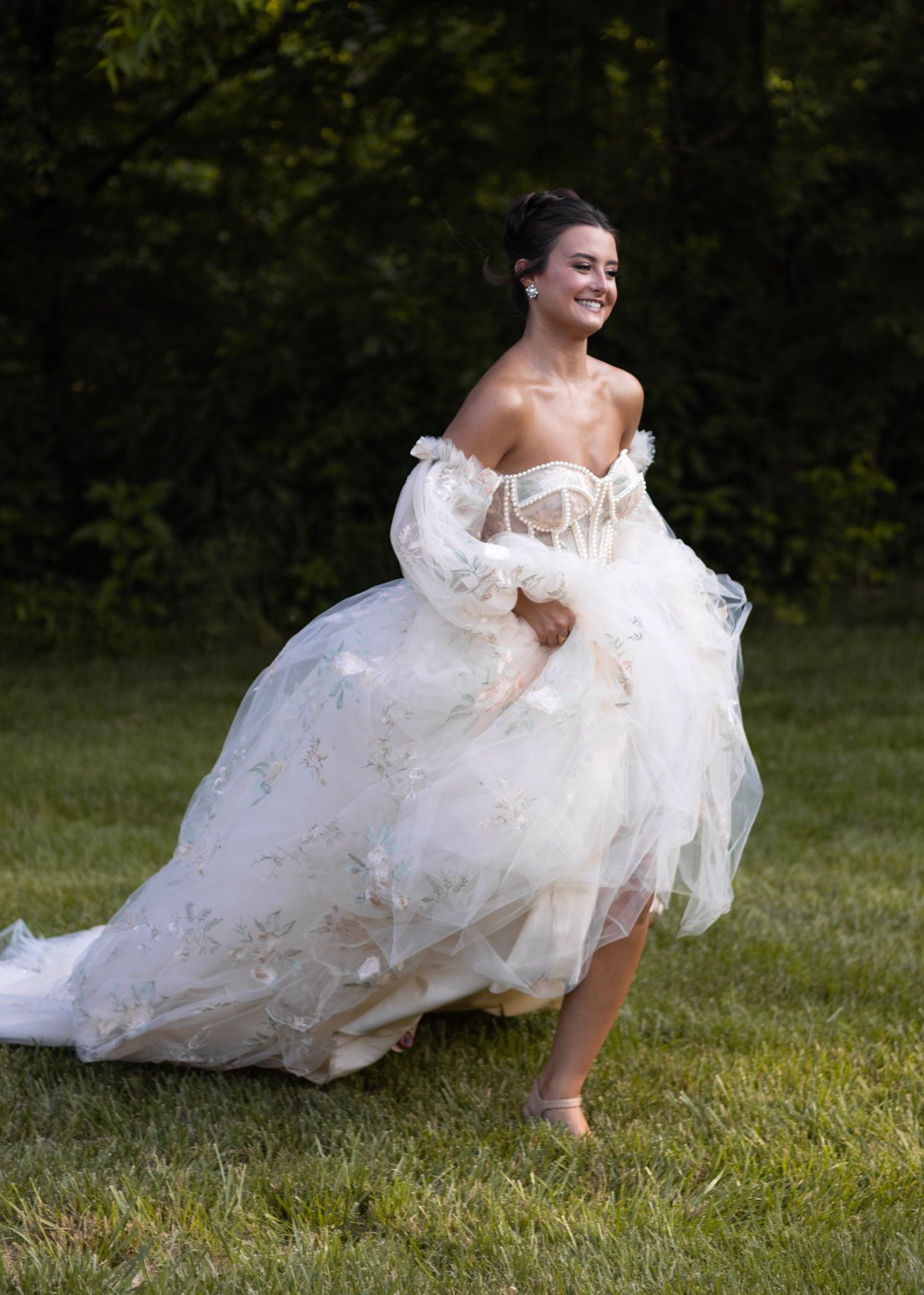 An ethereal off-shoulder wedding dress with flowing tulle skirt captured in a playful outdoor garden setting.
