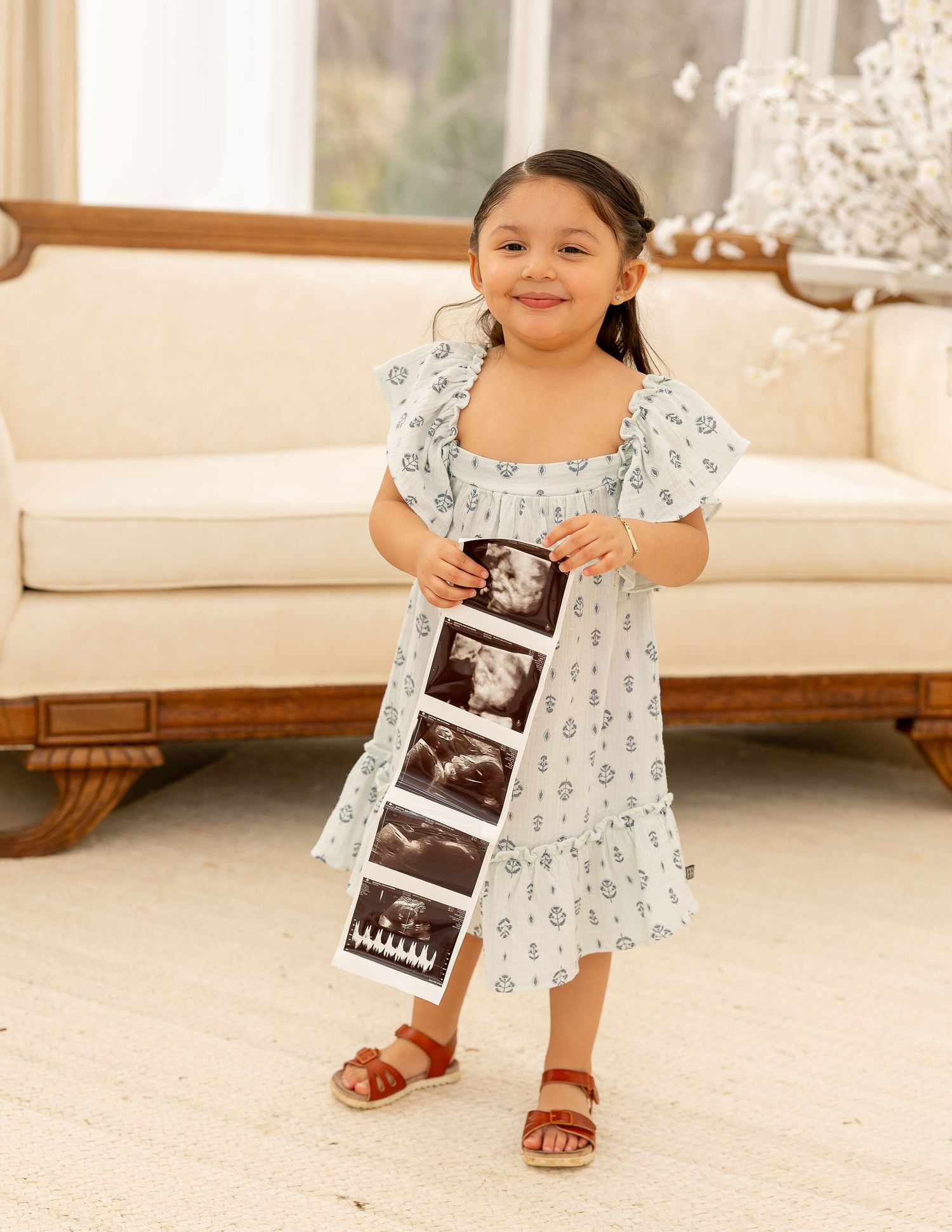 Young child in a floral dress holding ultrasound photos while sitting on a light wooden floor.