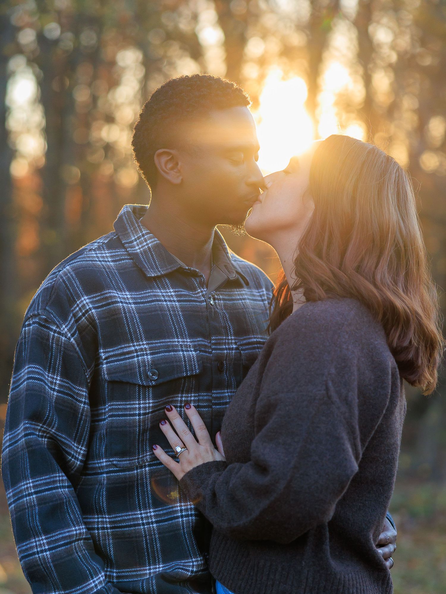 A couple embraces in a romantic outdoor photoshoot with golden sunlight filtering through trees in the background.