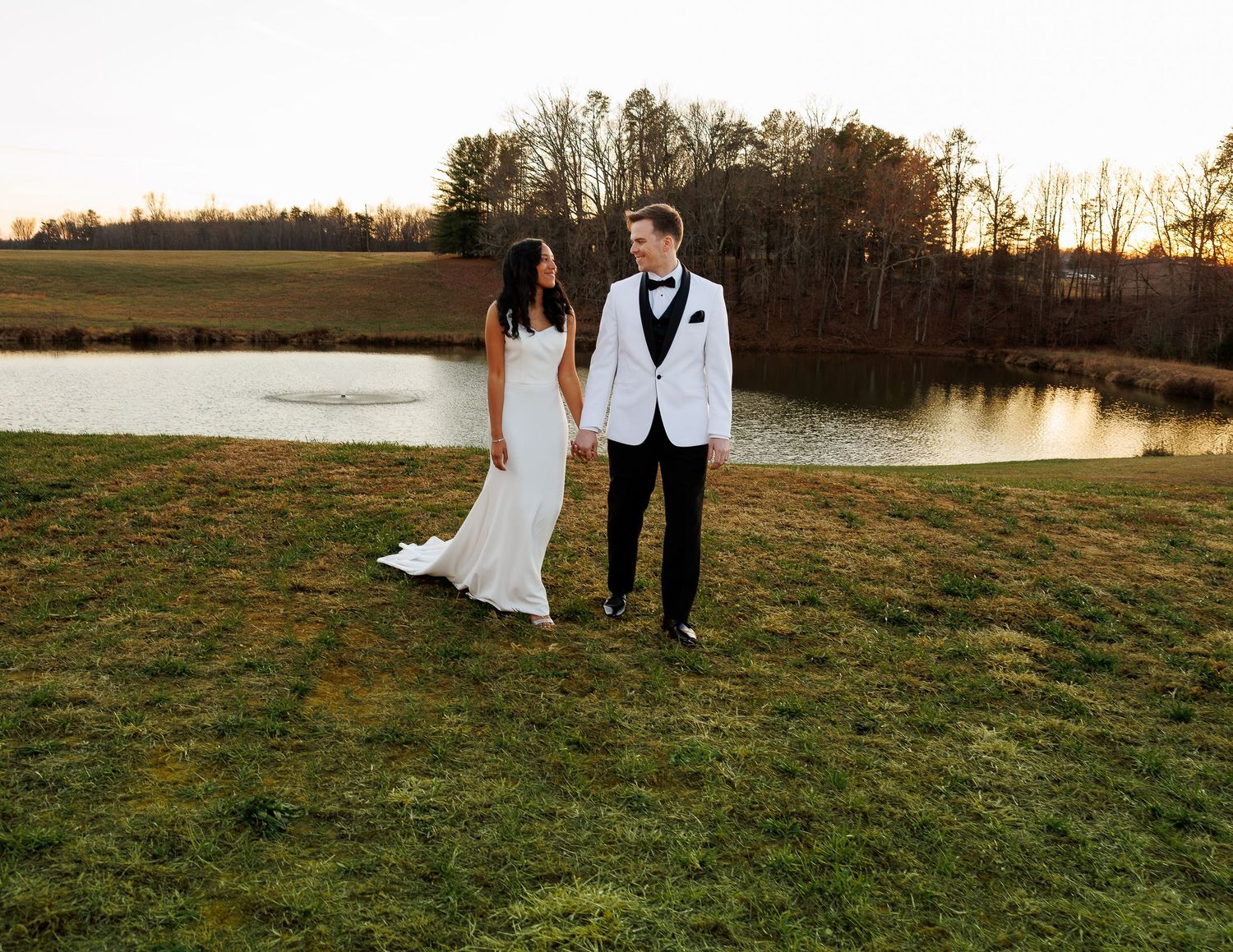A couple in formal wedding attire stands together on grassy lawn at sunset near a serene pond.