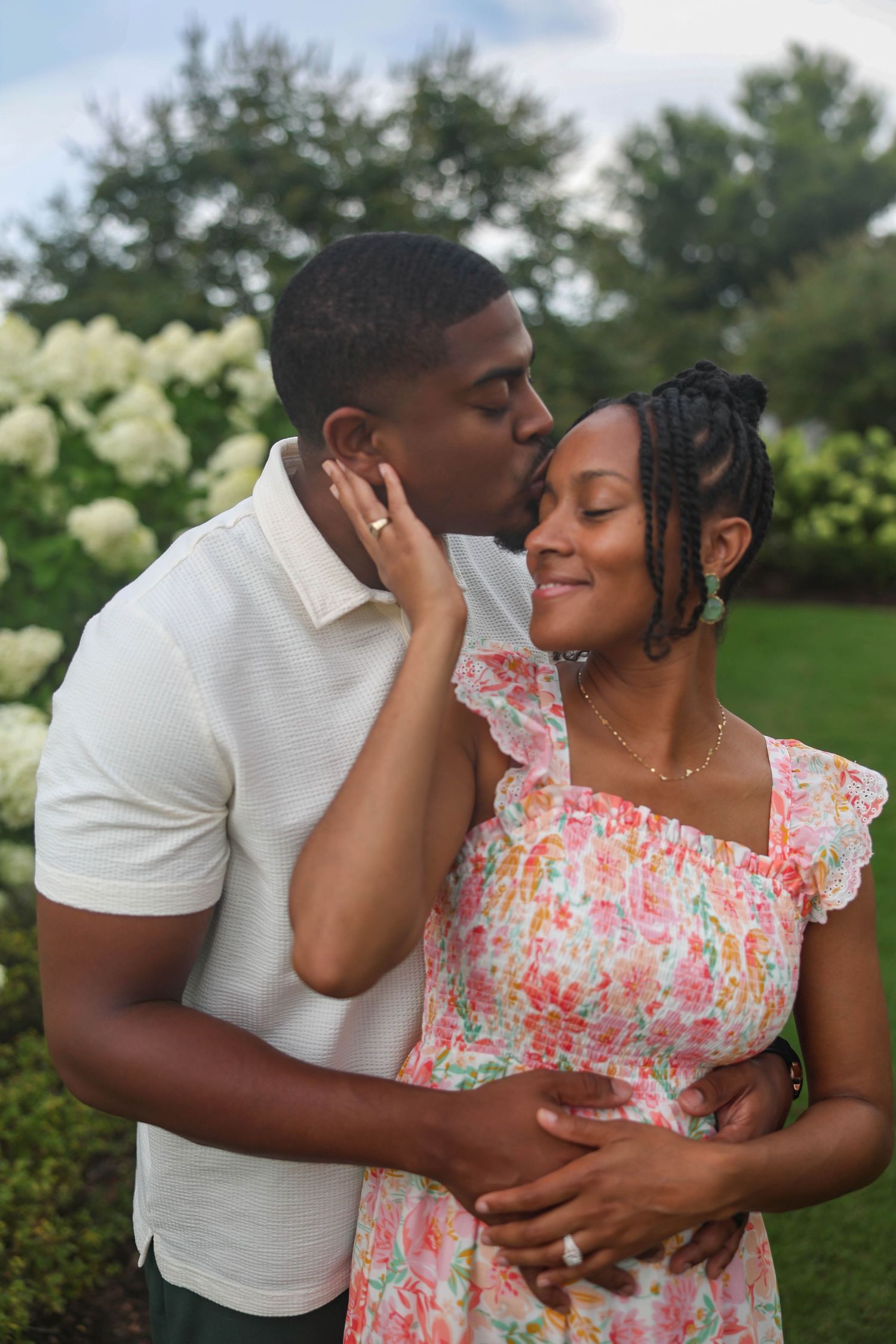 A couple shares a tender romantic moment in a garden with white flowers, with her wearing a floral pink dress.