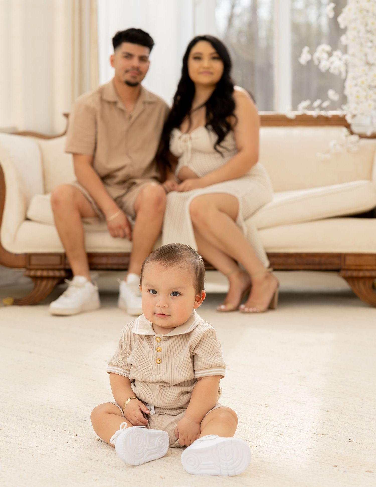 A baby in beige clothing sits on a light carpet while parents are visible in the background on a couch.