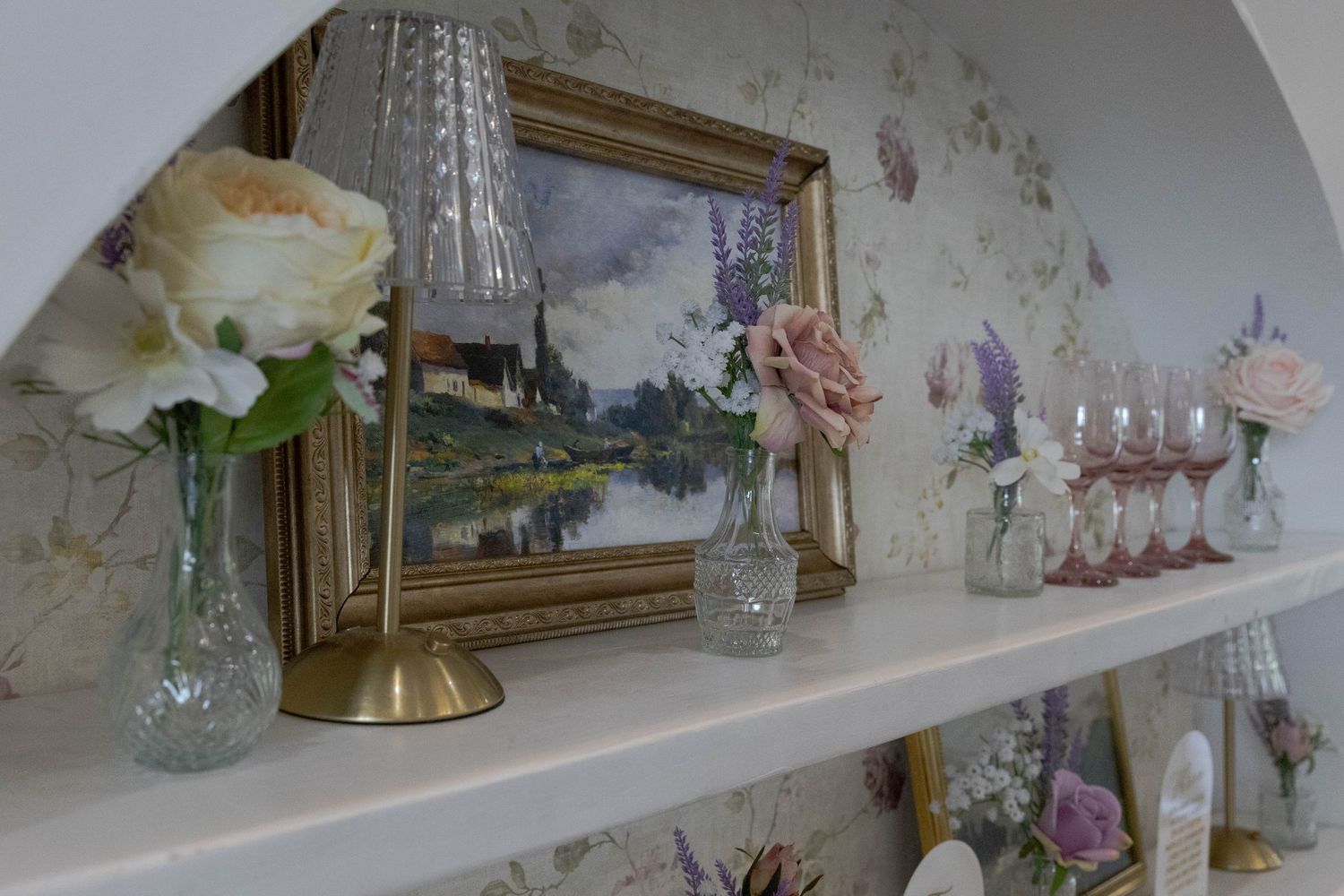 Vintage decor display with framed landscape painting, crystal glassware, and delicate floral arrangements on white shelf.