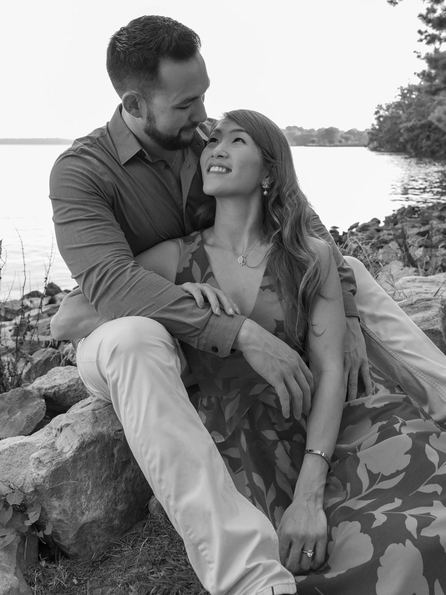 A couple in casual attire embraces romantically on rocky shoreline during sunset photoshoot by the water.
