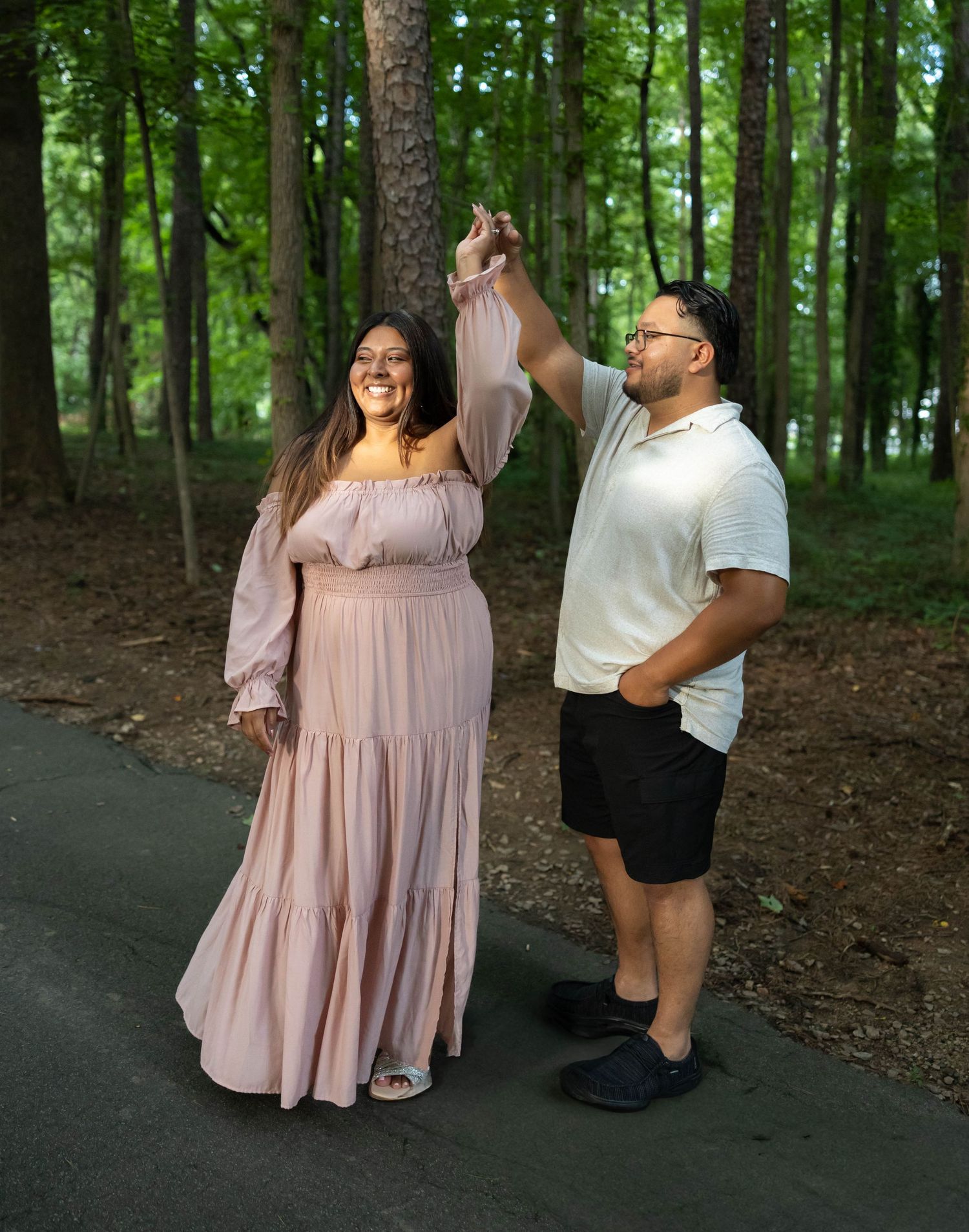 A couple stands together on a path in the woods, wearing casual clothing with the person on the right raising their arm up.