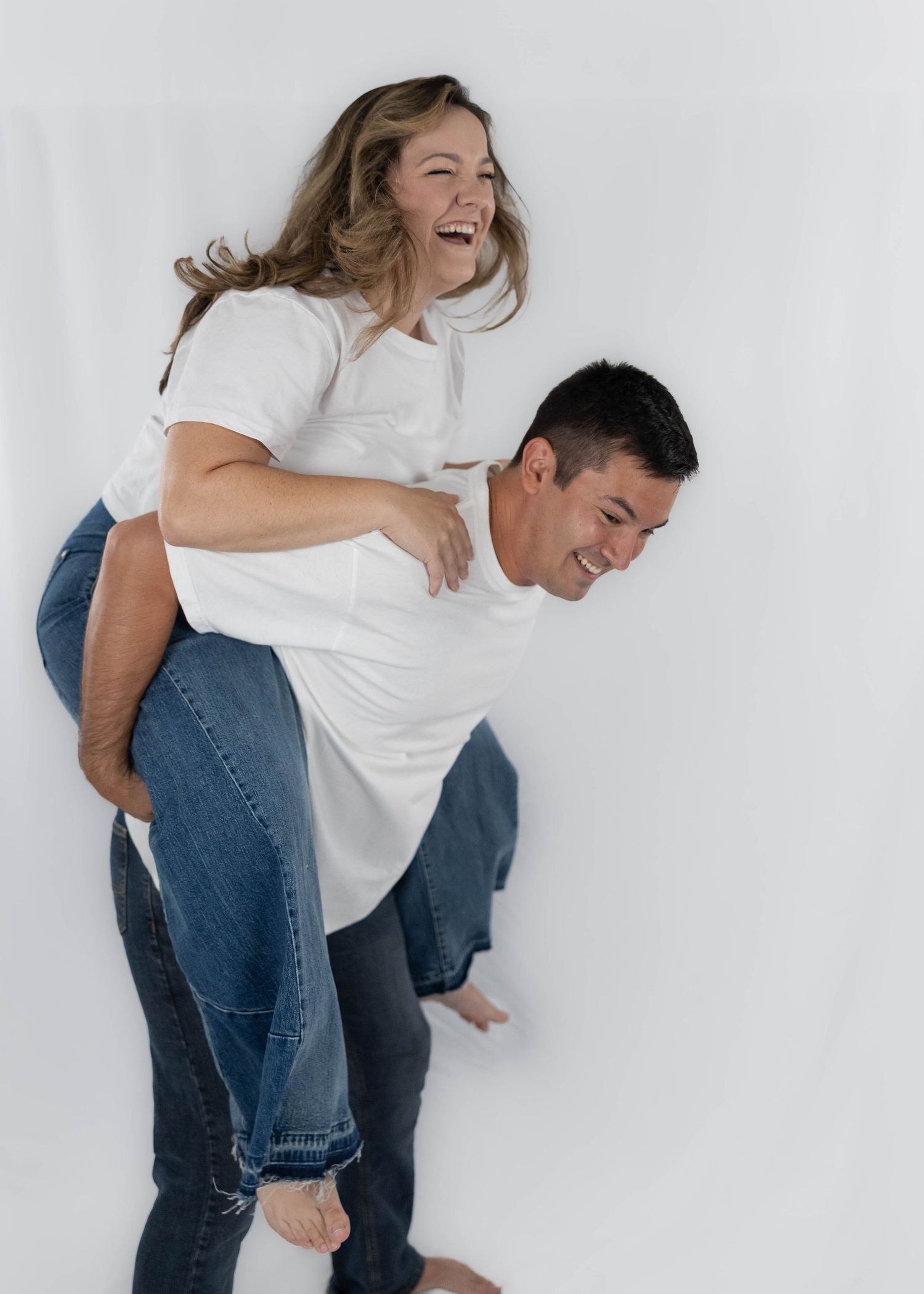 A playful couple in white t-shirts and jeans laugh together against a white backdrop during a casual photoshoot.