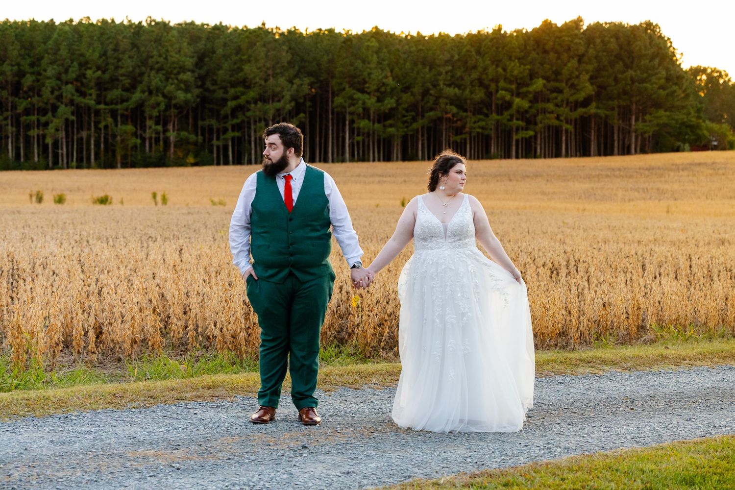 Bride in white gown and groom in green suit hold hands walking on gravel path beside golden field at sunset.