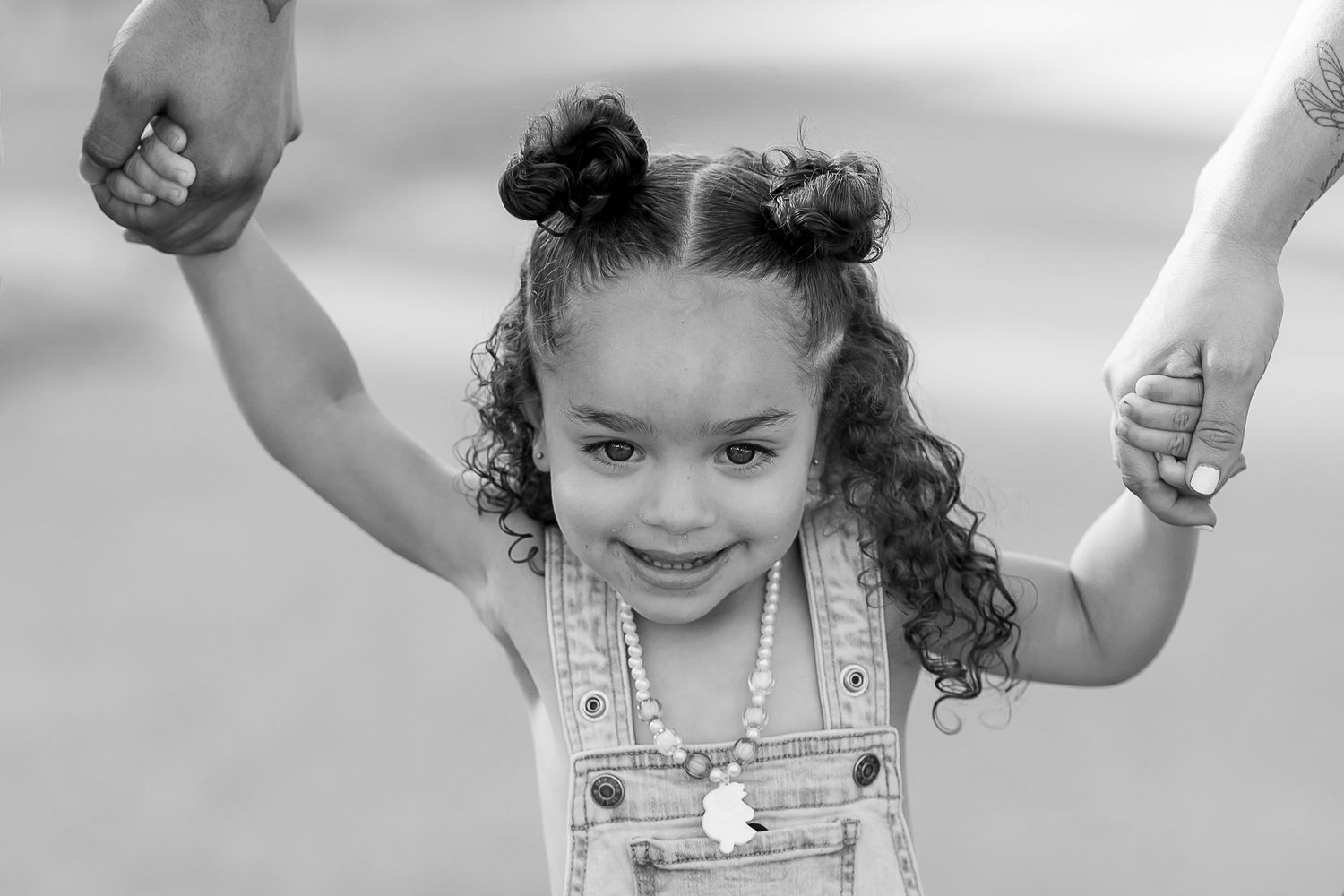 Adorable little girl with curly hair and bun pigtails holding parents hands in black and white photo.