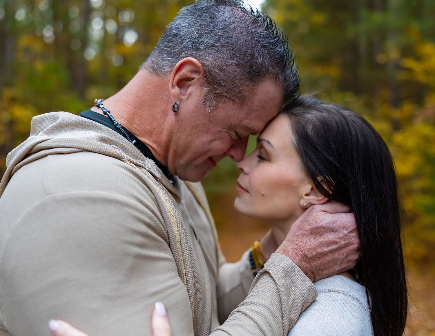 A romantic couple embraces and kisses in a park with autumn foliage in the background.
