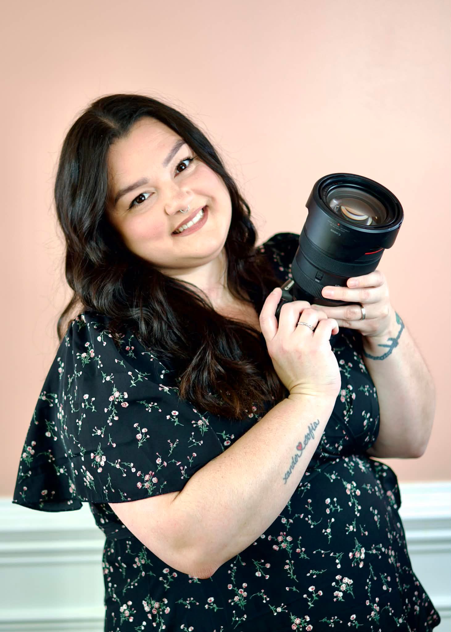 A photographer in a floral dress demonstrates different playful poses while holding a camera against a pink background.