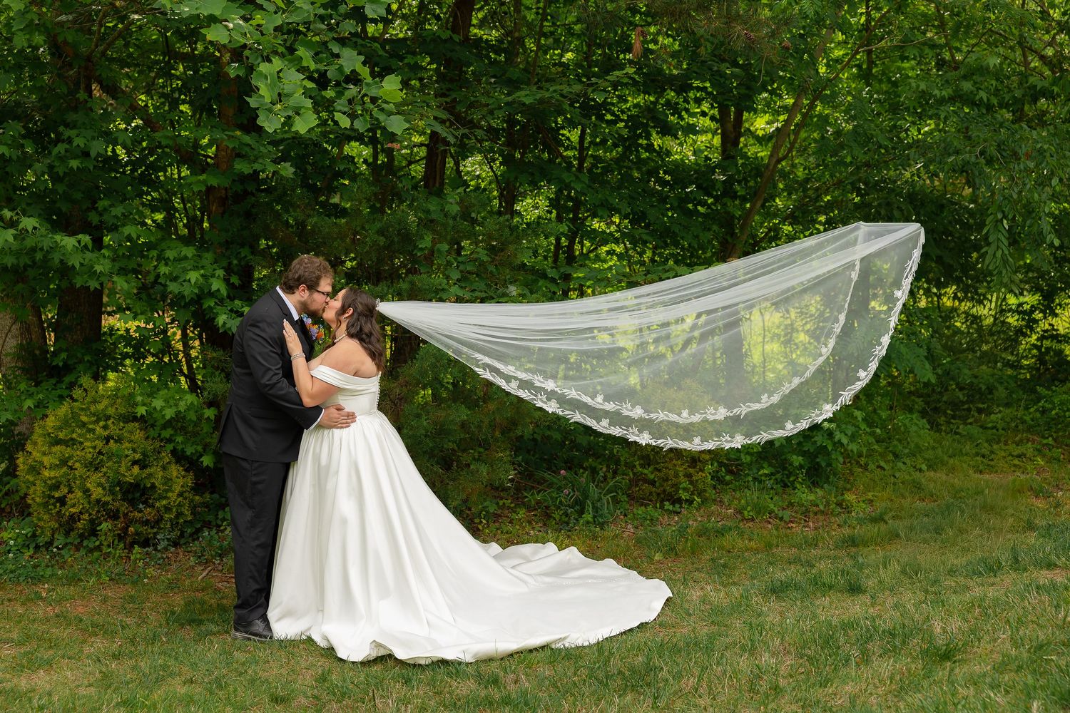 Bride and groom share a romantic kiss outdoors with wedding veil flowing in the wind against lush green trees.