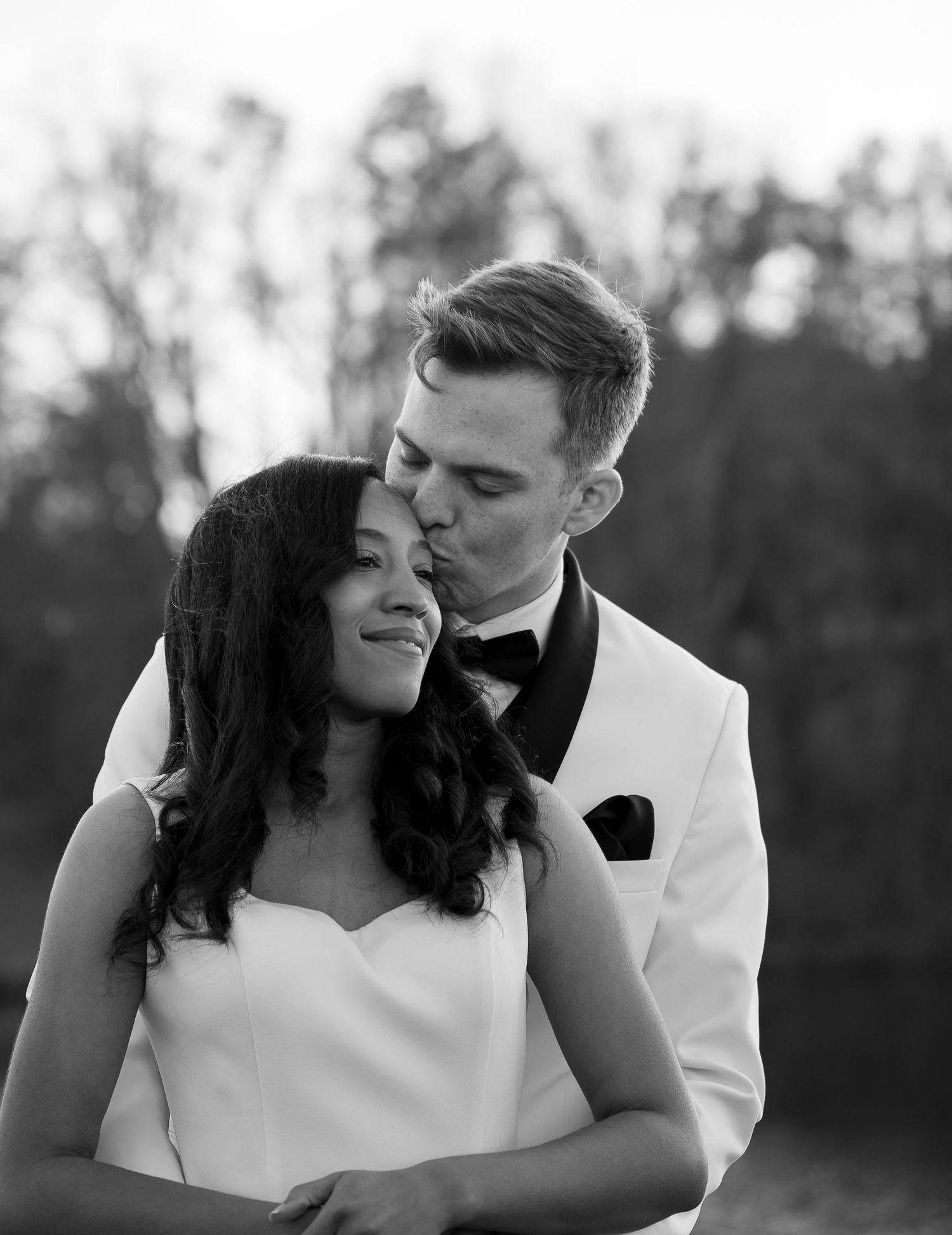 Black and white wedding photo of groom in white tuxedo kissing bride on forehead outdoors.