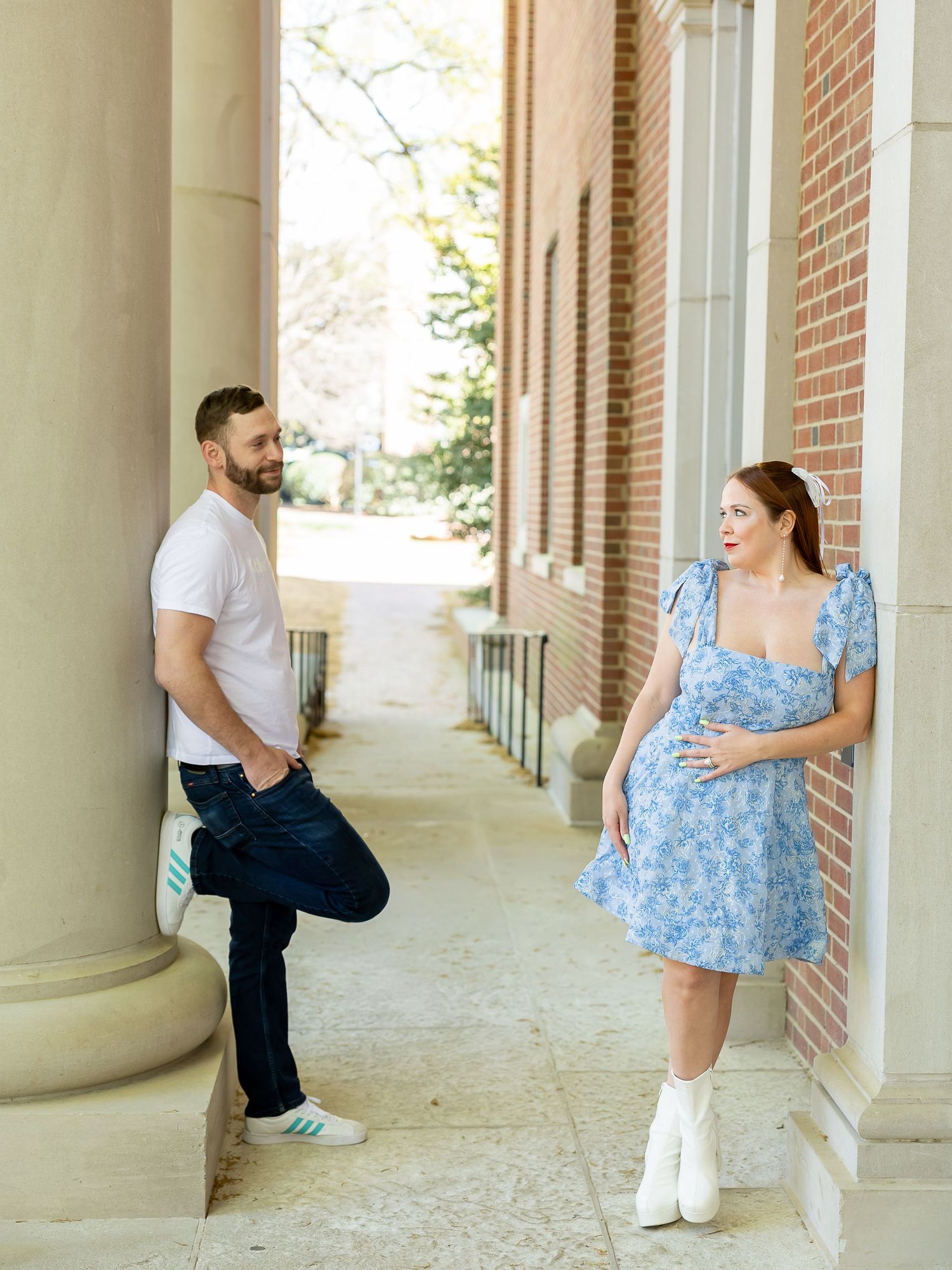 A pregnant woman in a blue floral dress and a man in a white shirt and jeans stand together in a brick-lined walkway.